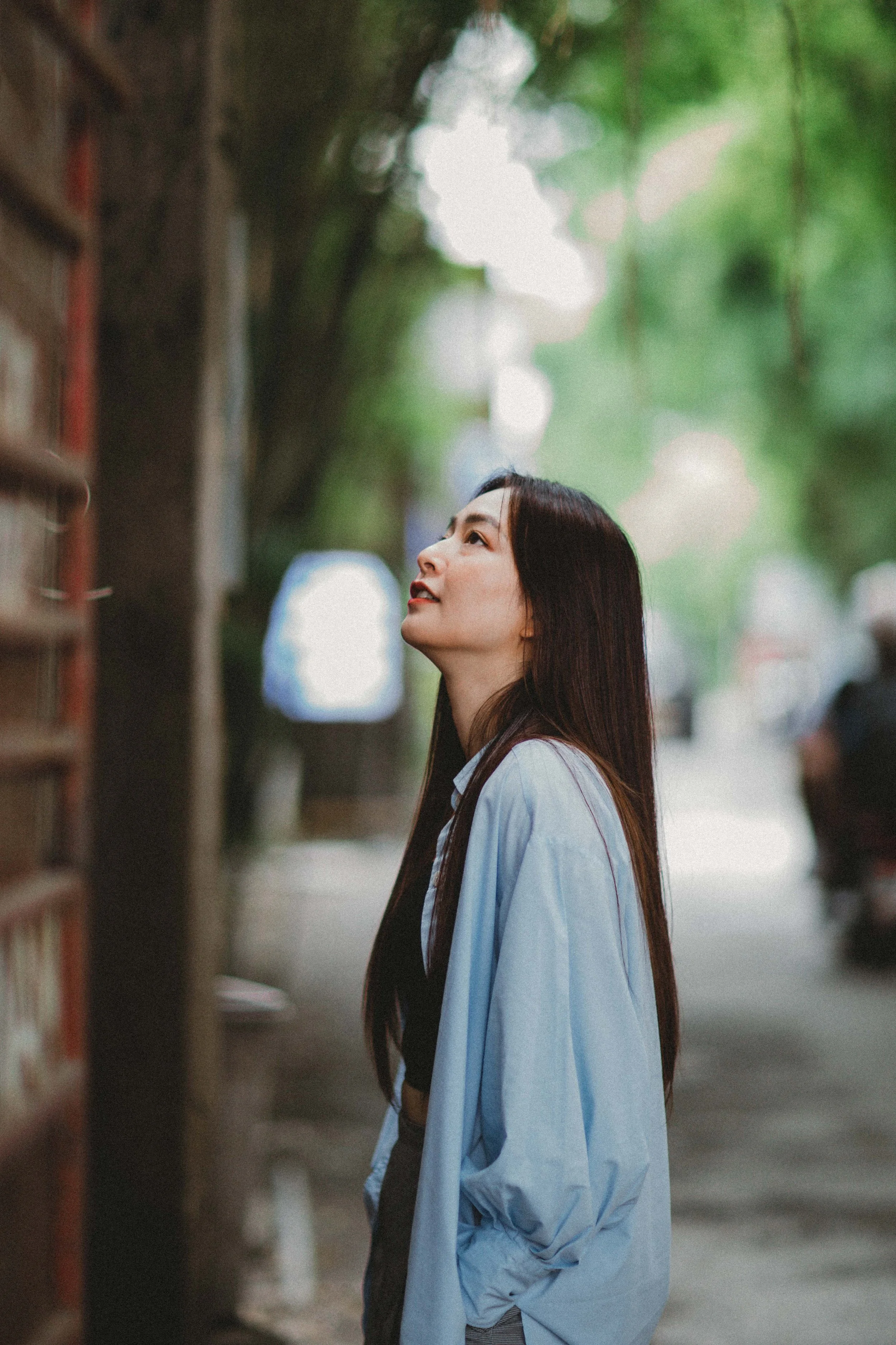 A woman with long dark hair wearing a light blue shirt looks upward along a wooded street.