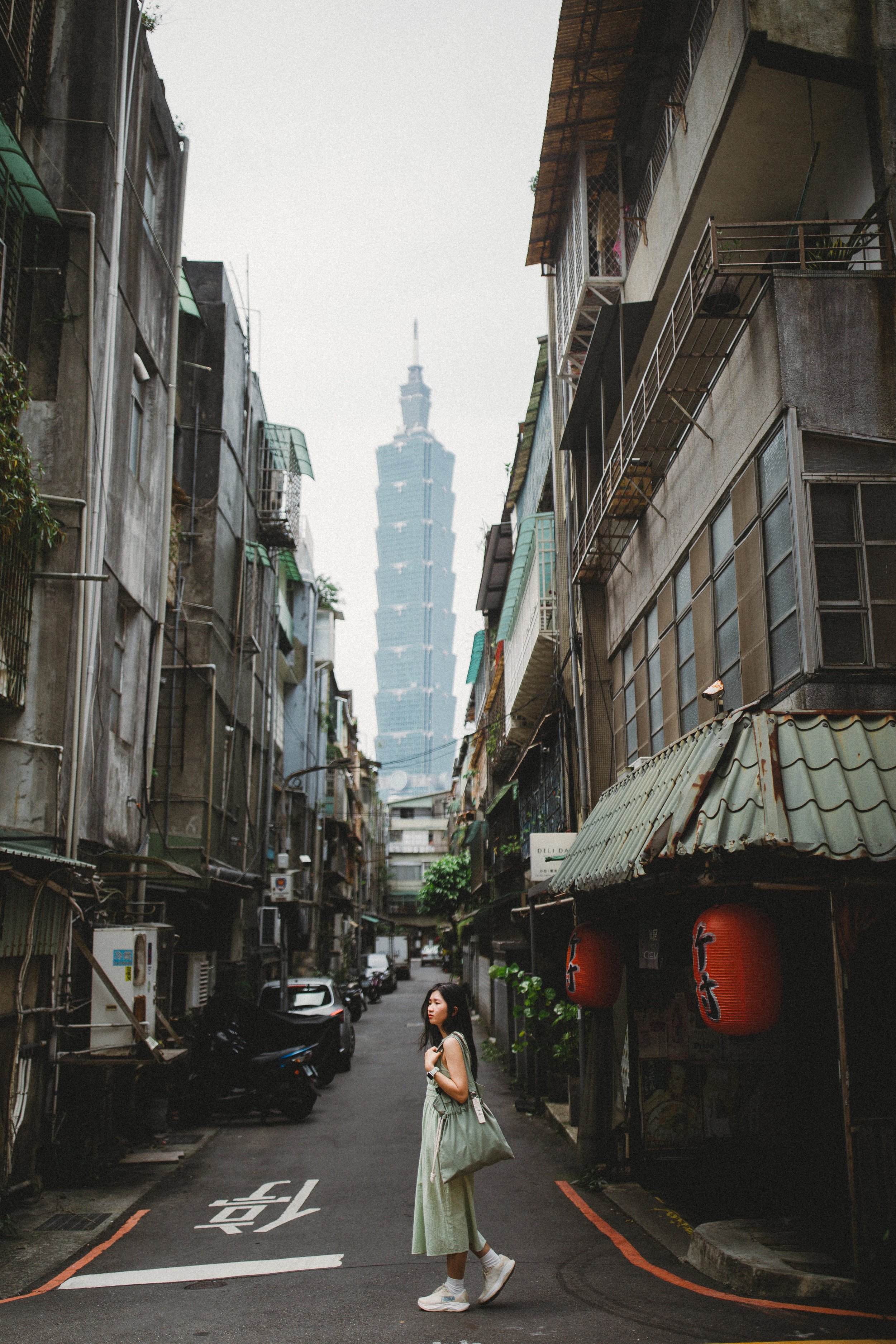 A young woman with a backpack and large bag walks through a narrow urban alley with old buildings, and Taipei 101 skyline in the background.