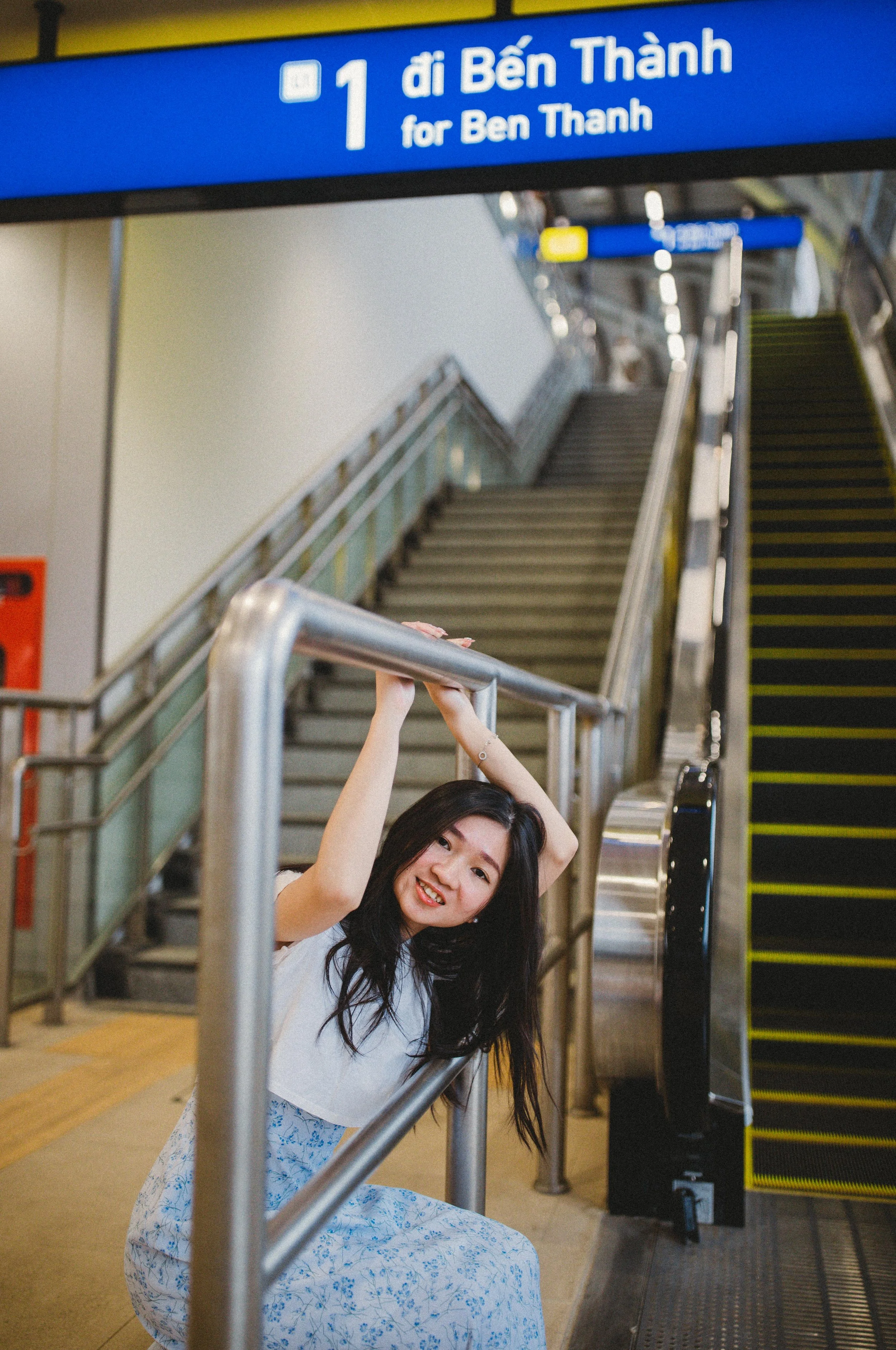 A young woman with long dark hair smiling and sitting on the floor at an airport terminal, leaning against a metal railing near an escalator, with a sign above indicating direction to Ben Thanh.