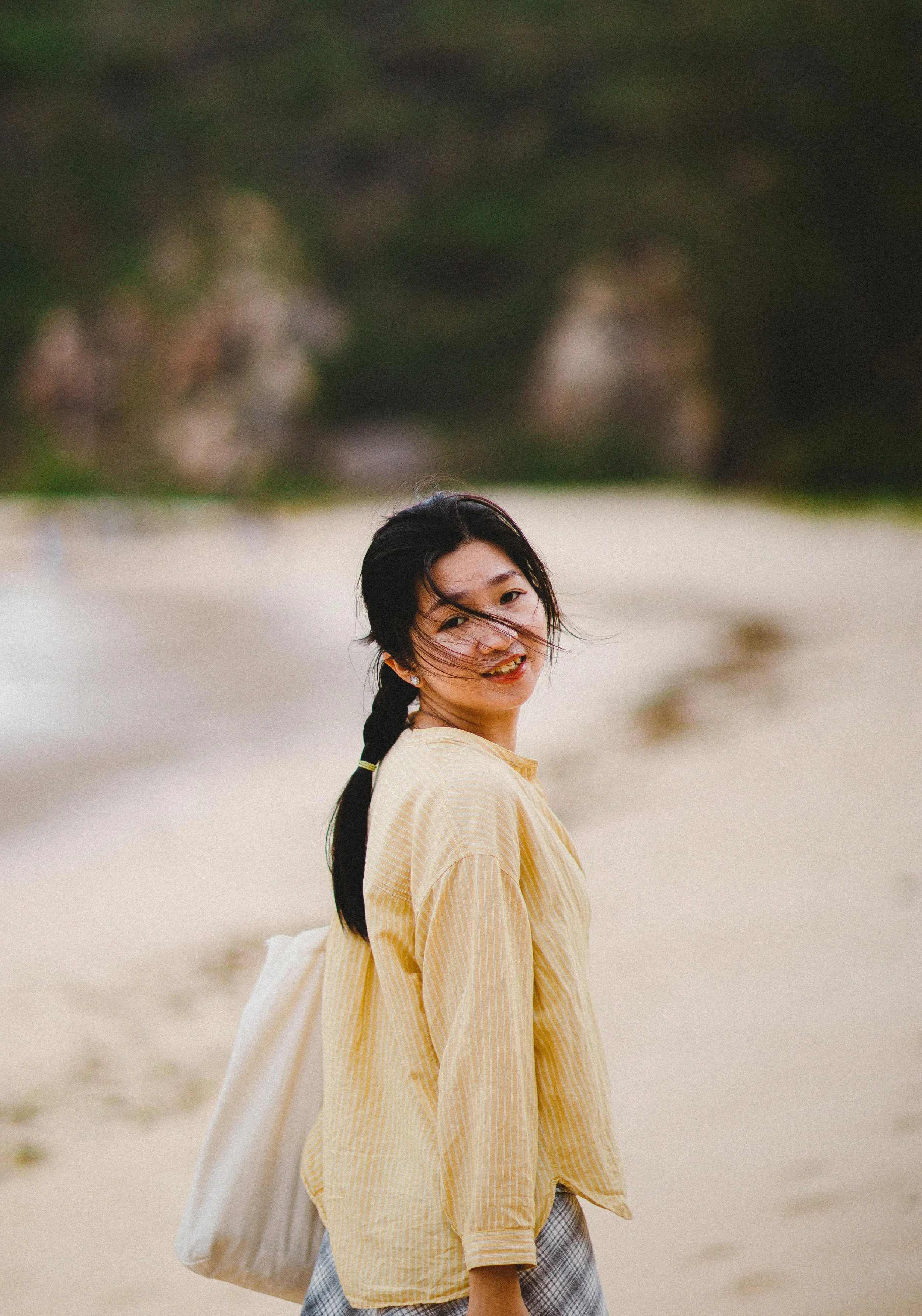 Young woman with long dark hair in a braid, smiling and looking at the camera on a sandy beach with trees in the background.