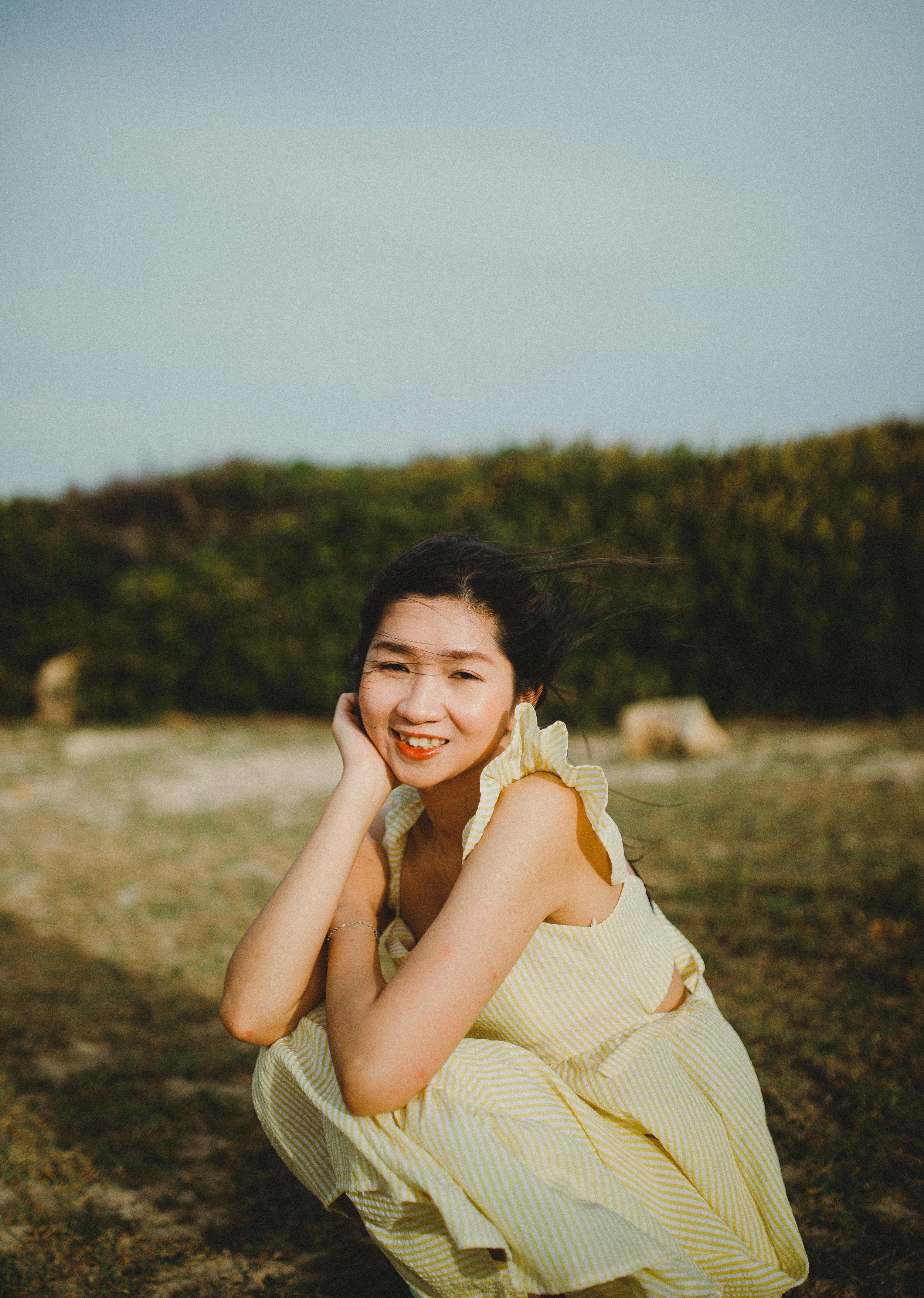 A woman in a yellow and white striped dress squats outdoors, smiling and touching her face with her right hand, with windblown hair and a field in the background.