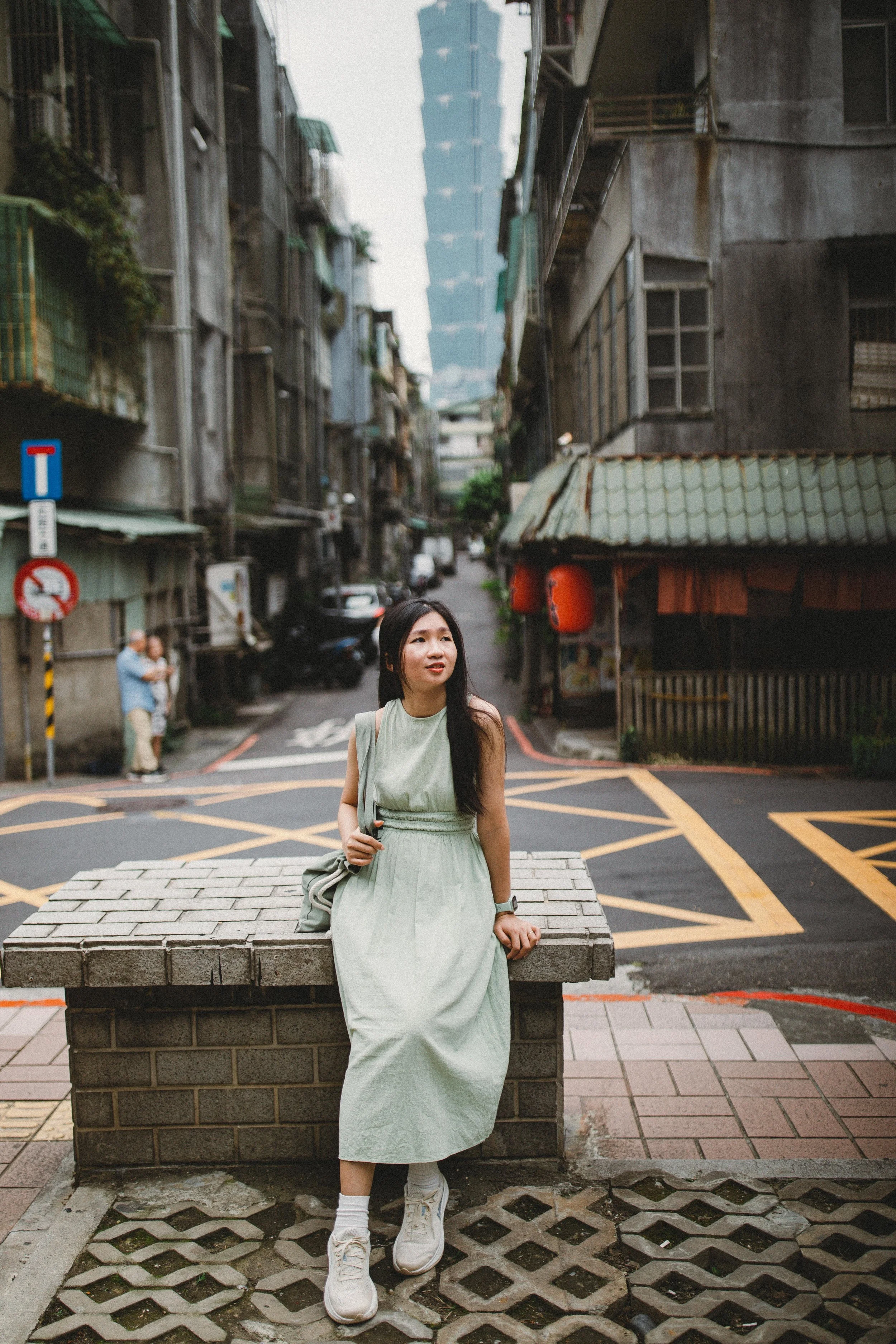 A woman in a light green dress sitting on a brick bench in an urban street scene with tall buildings and a modern skyscraper in the background.