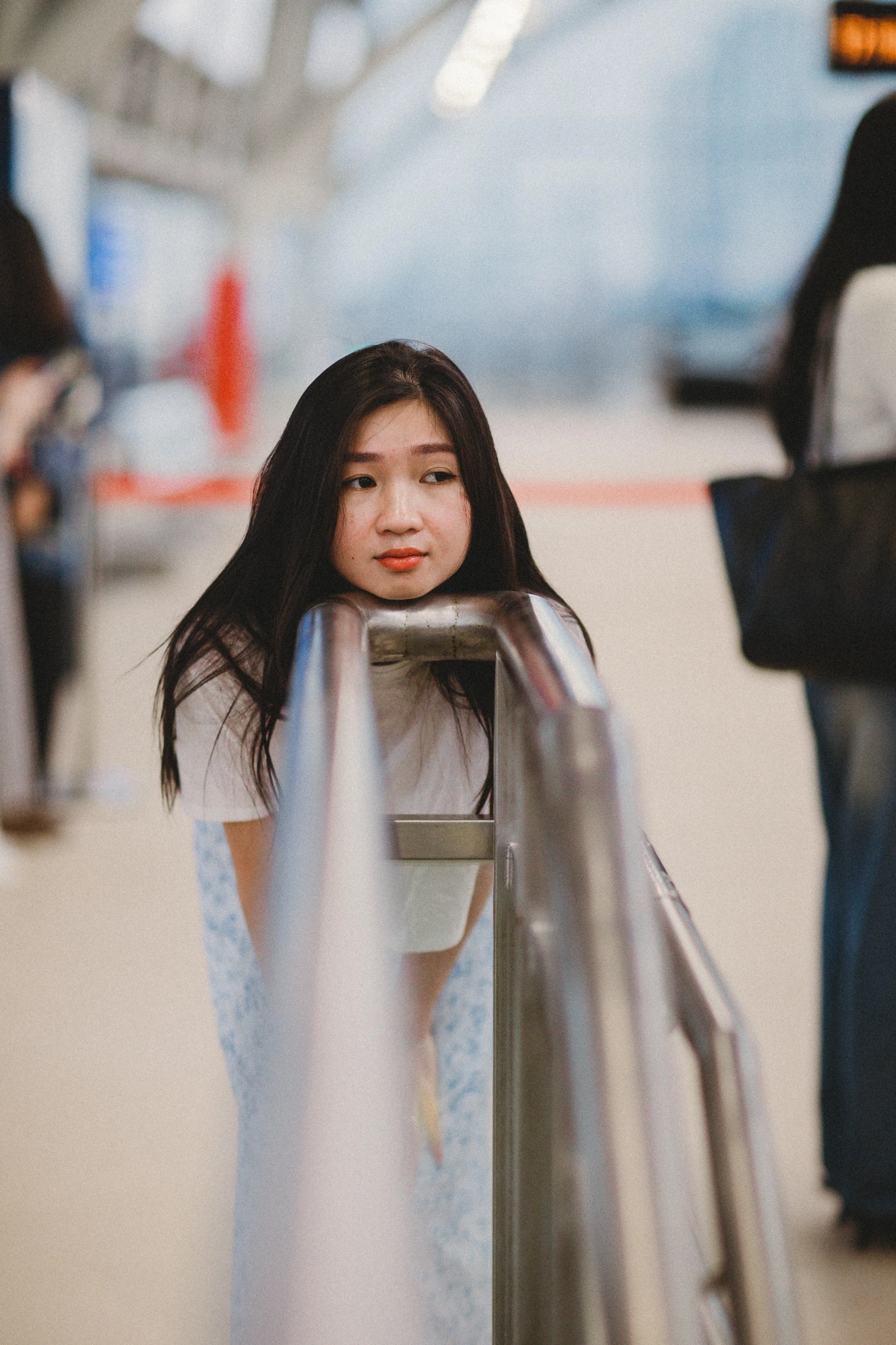 A young woman with long dark hair leaning on a metal railing at an airport, looking contemplative.