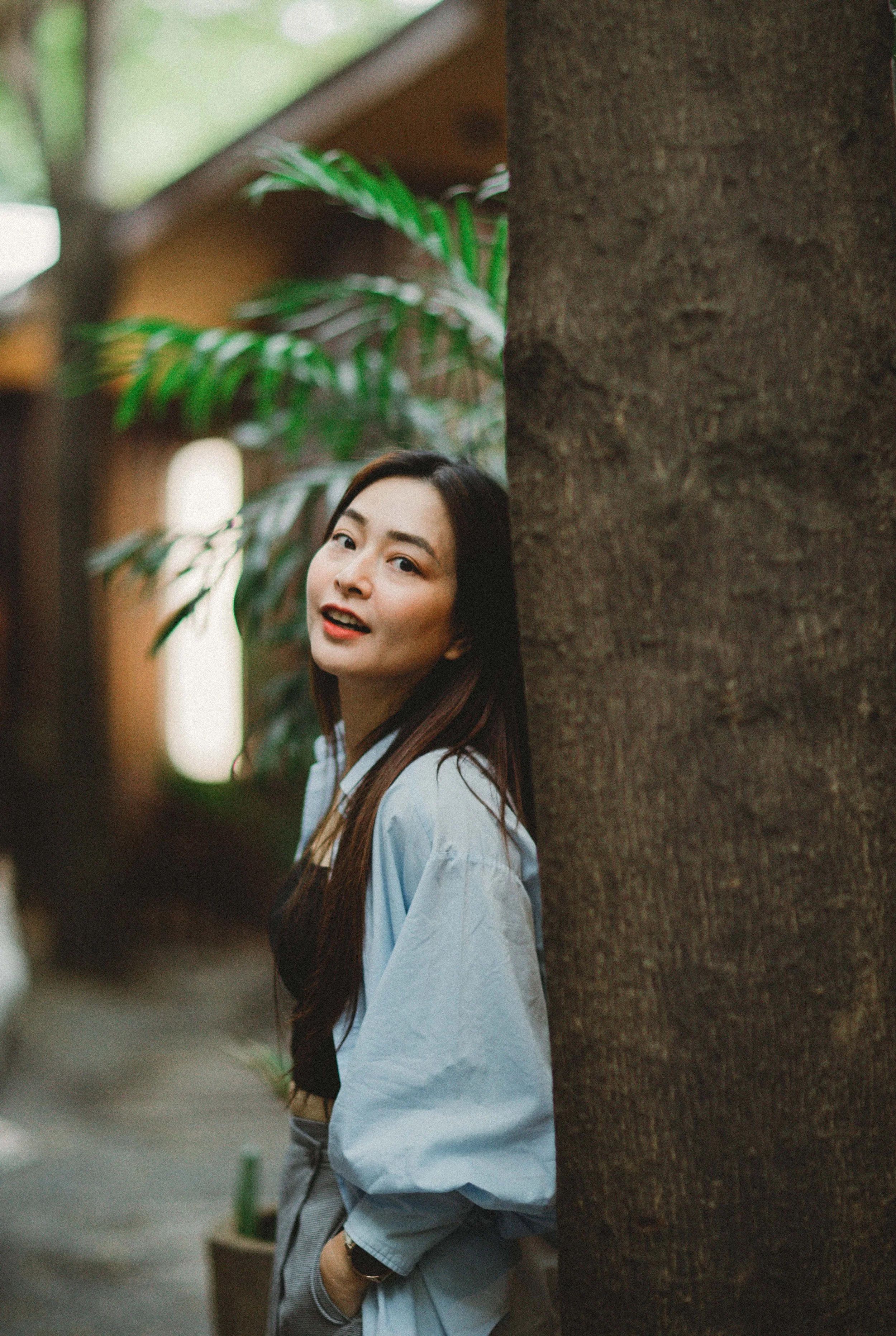 A woman with long dark hair leaning against a tree, looking at the camera, in an indoor space with greenery and wooden elements.