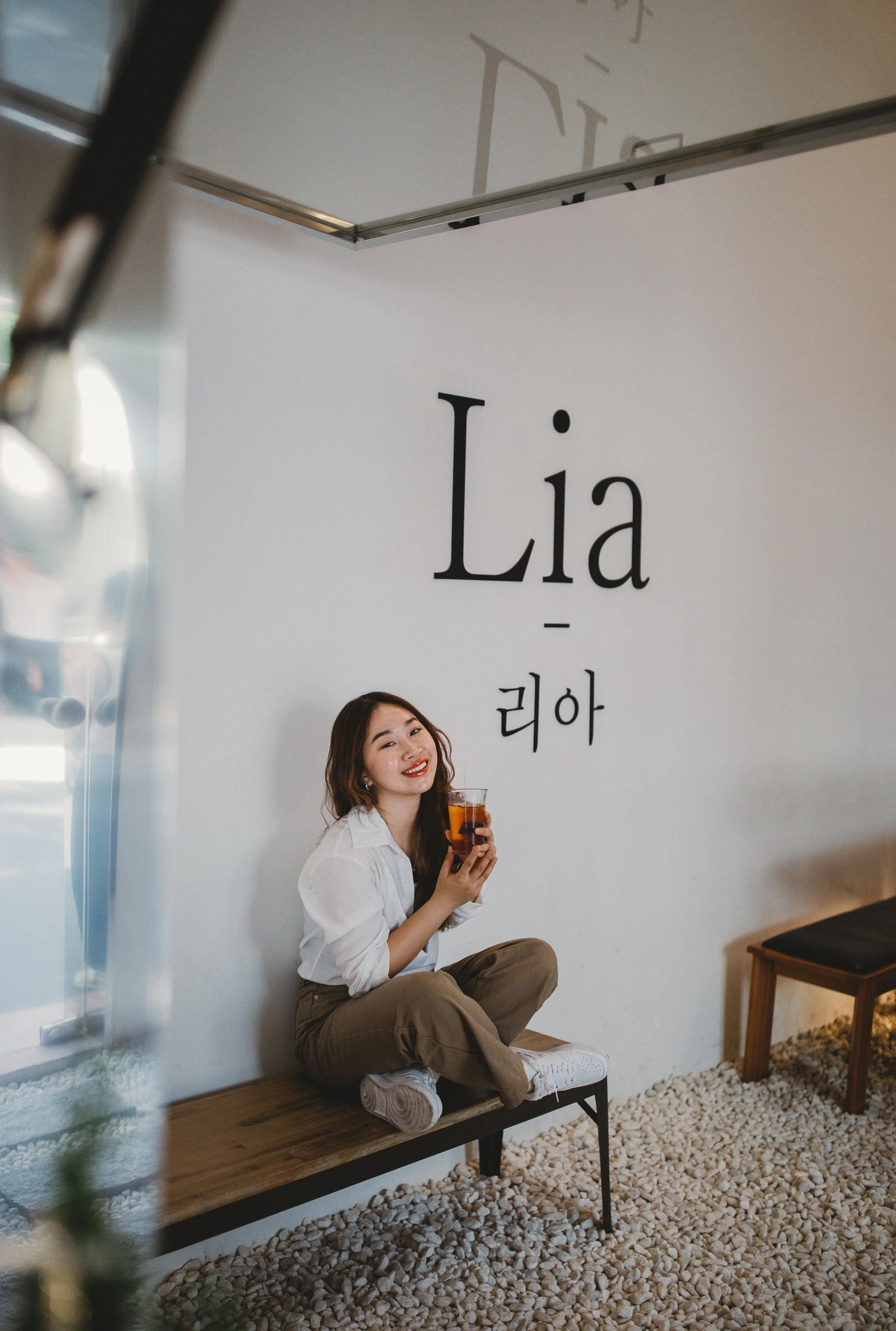 A young woman with long dark hair, sitting cross-legged on a wooden bench inside a cafe, holding a glass of iced drink, smiling at the camera. Behind her is a white wall with the word 'Lia' written, along with Korean characters.