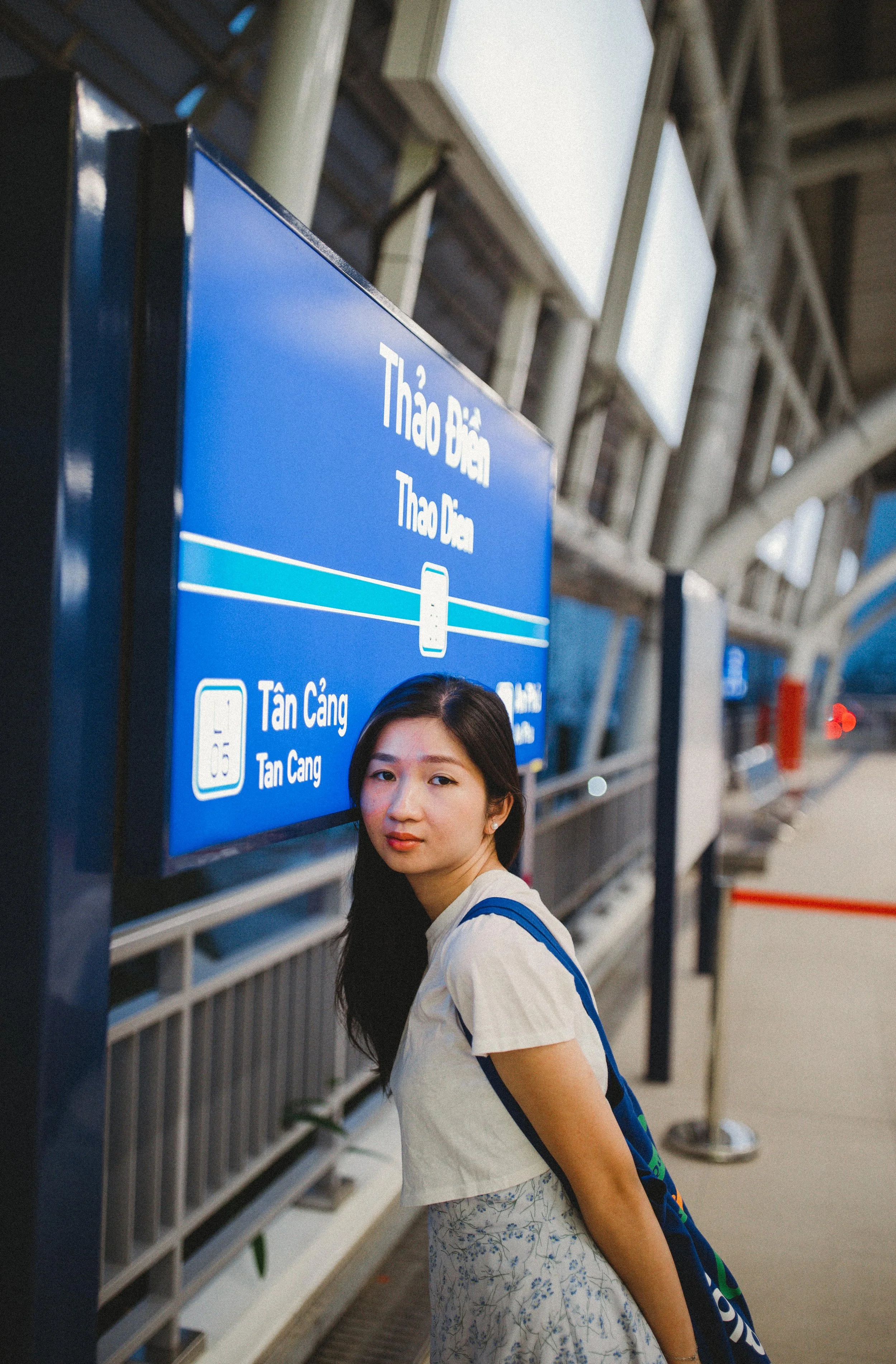 A young woman with long black hair standing at a transit station near a blue sign with white text. The sign displays the station name 'Thao Dien' in both Vietnamese and English, with an additional note 'Tan Cang,' and a graphic of a train or metro wi