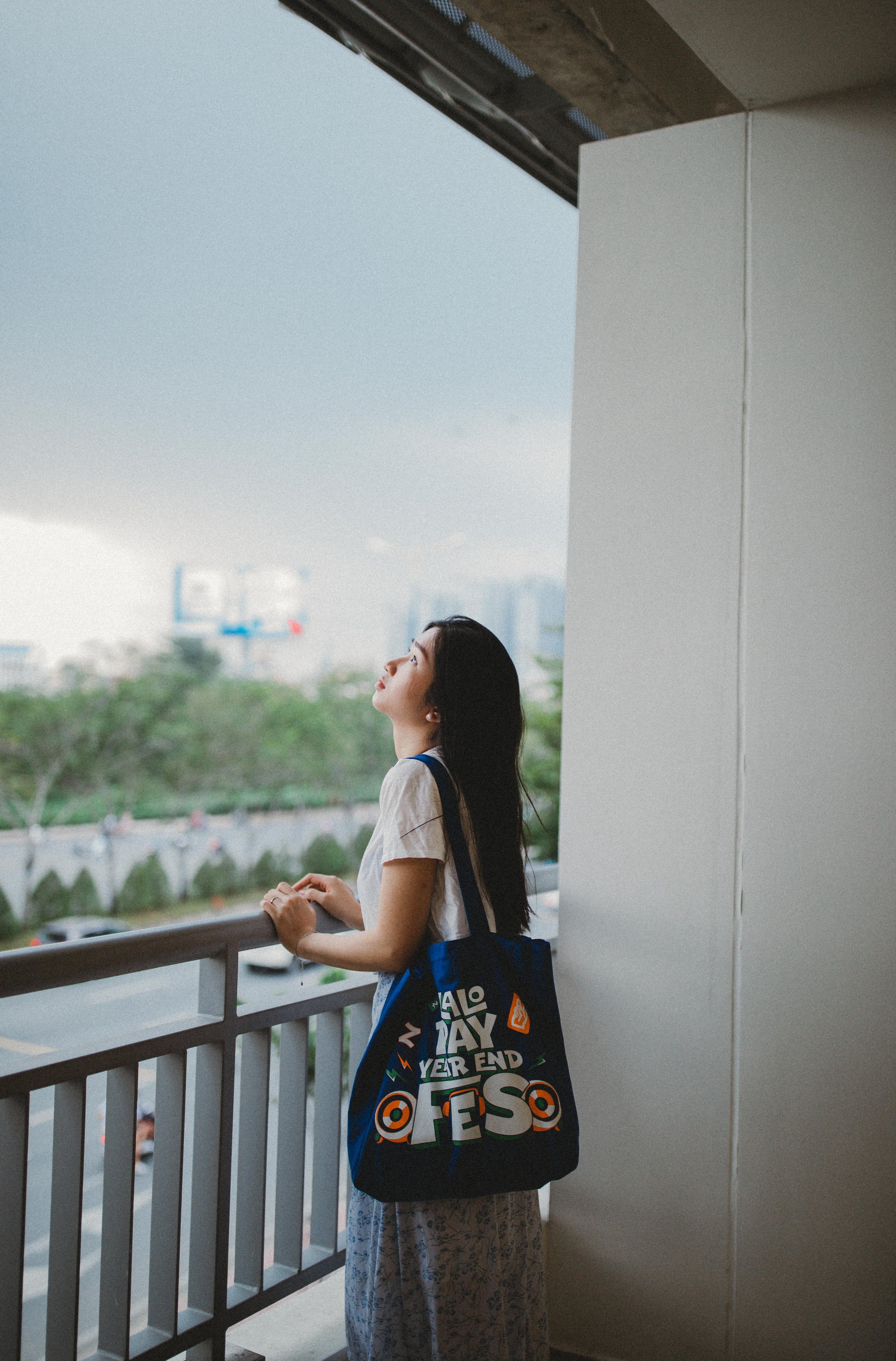 A young woman with long dark hair stands on a balcony, gazing upward with her eyes closed, holding onto the railing. She wears a white T-shirt, patterned pants, and carries a large dark blue tote bag with colorful text and graphics. The background fe