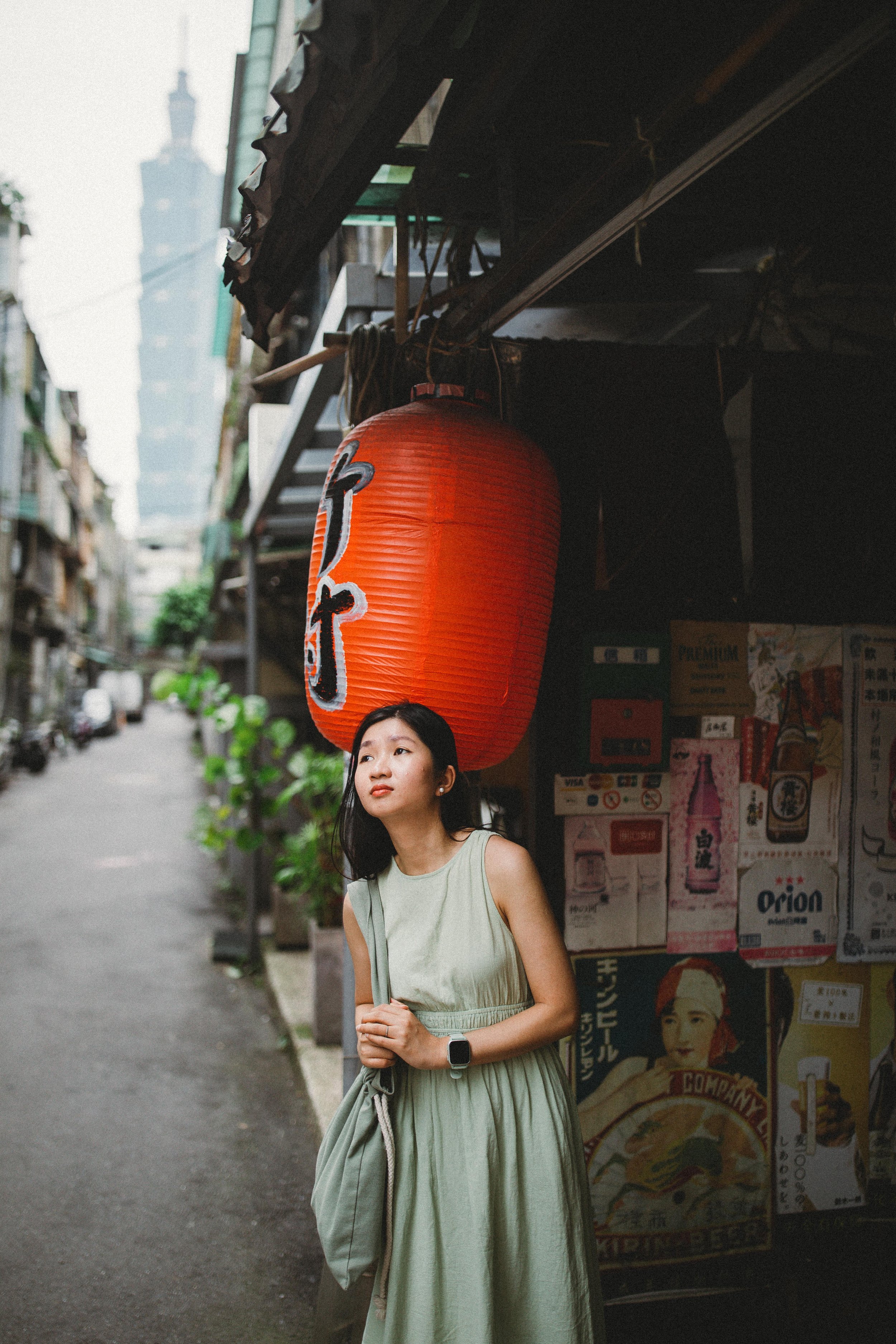 A young woman in a light green dress stands on a street in front of a food stall with a large red paper lantern and Asian posters. Tall building in Taipei 101 tower is visible in the background.