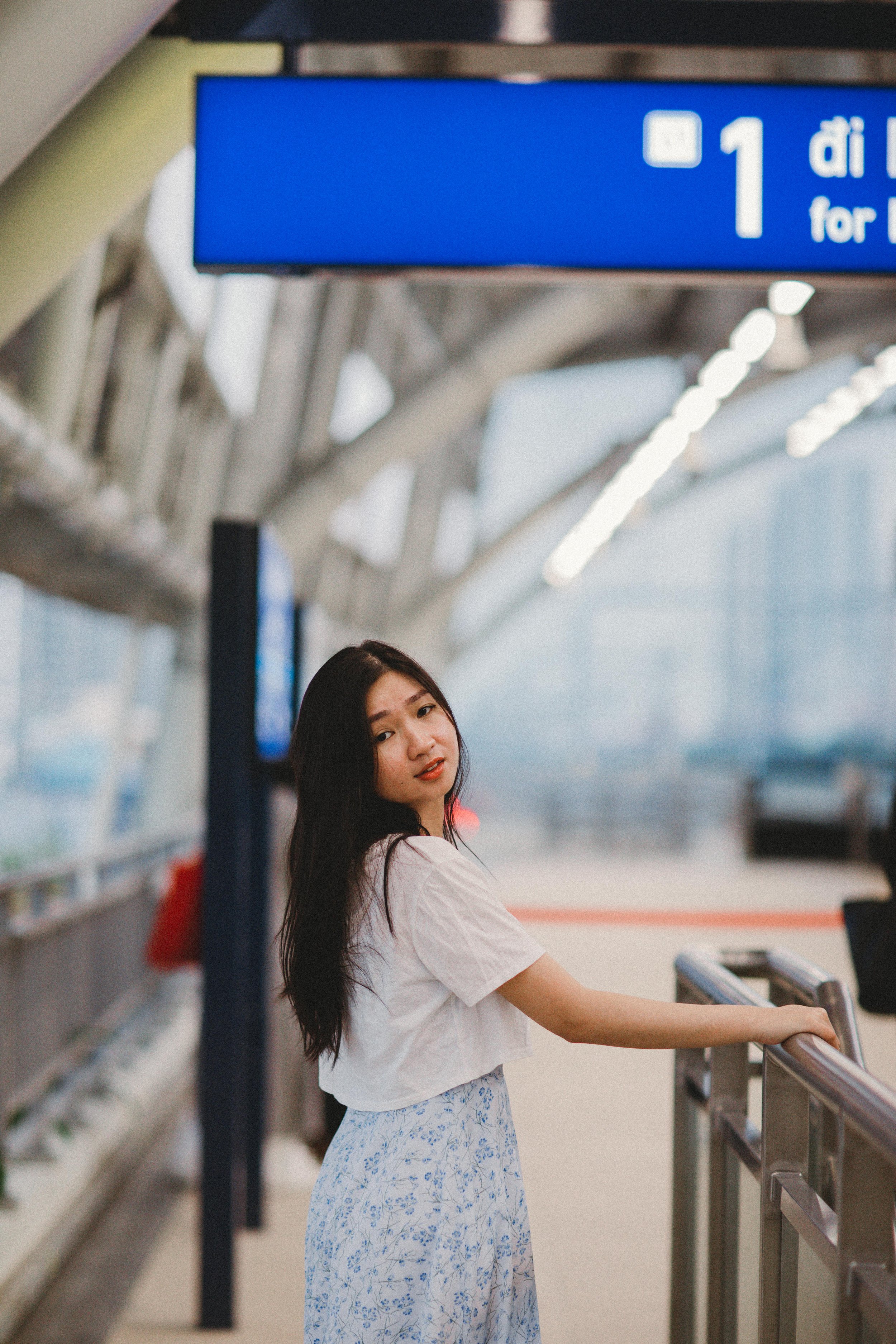 A young woman with long dark hair standing at an airport terminal, holding onto a railing with her right hand, with blue signage above her indicating direction for Gate 1.
