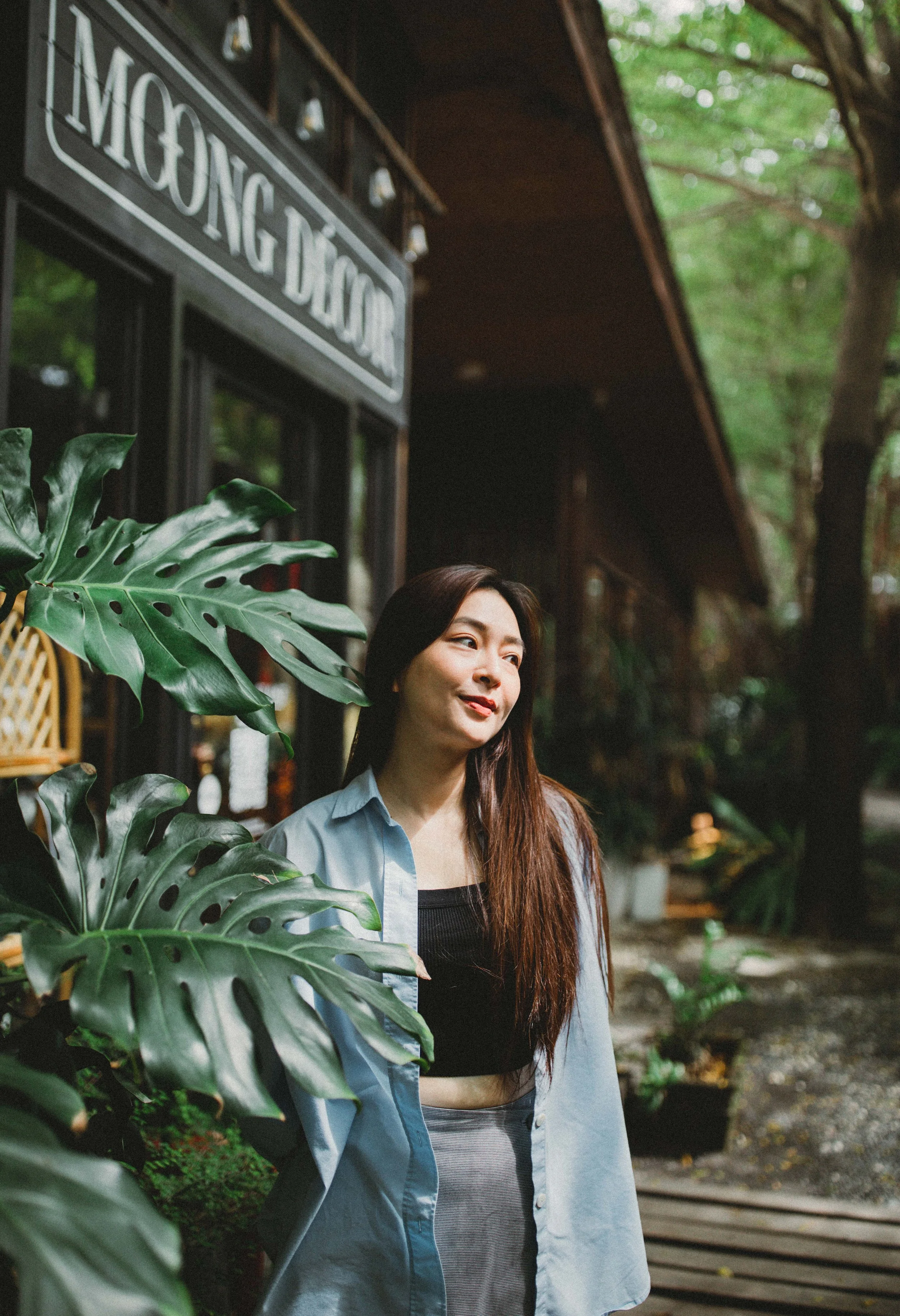 A young woman with long brown hair standing outside a shop called "MONG DOG" surrounded by large green tropical leaves, in a lush, wooded area.
