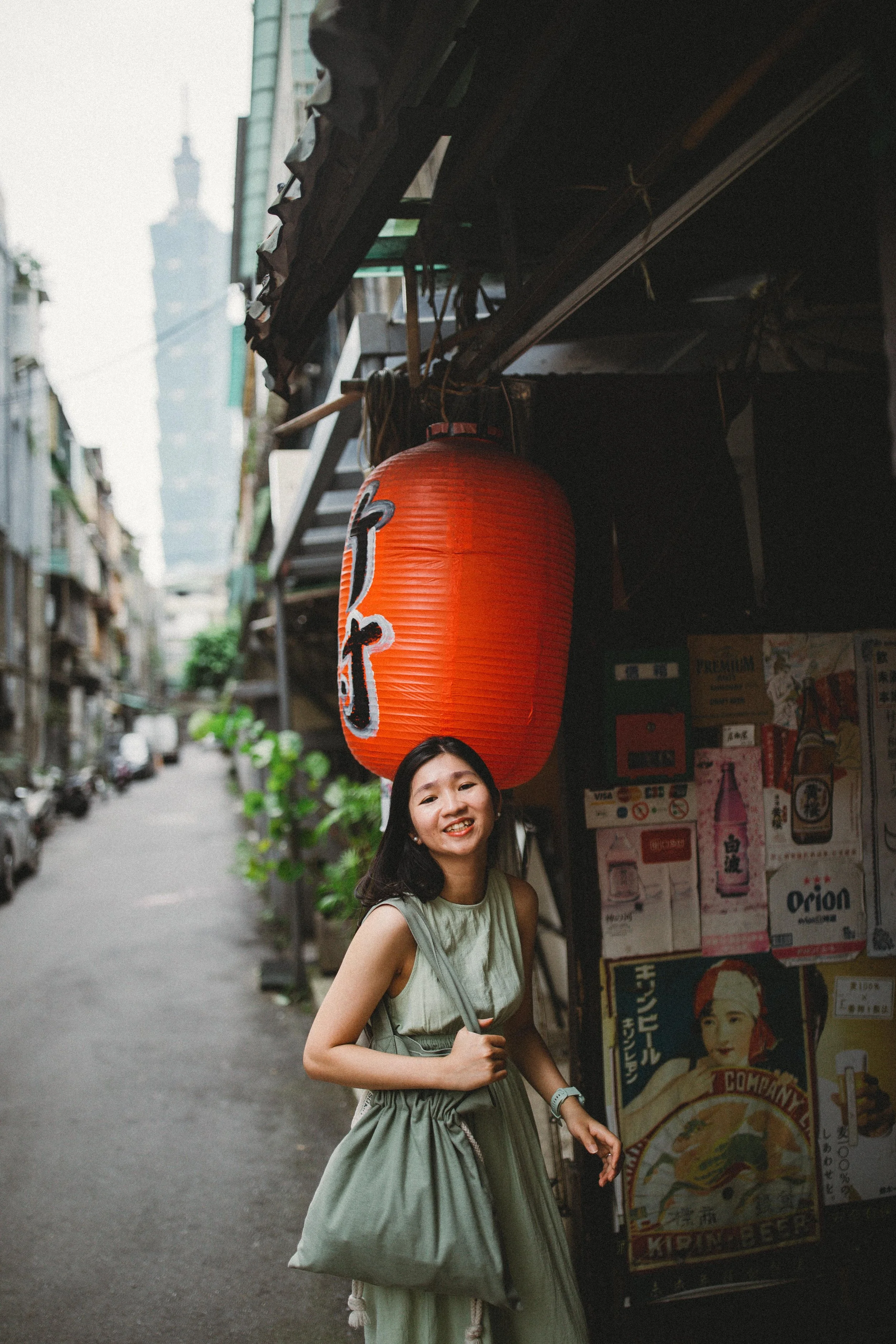 Young woman in a green dress smiling in an alleyway, standing near a red paper lantern and a wall with Japanese posters.