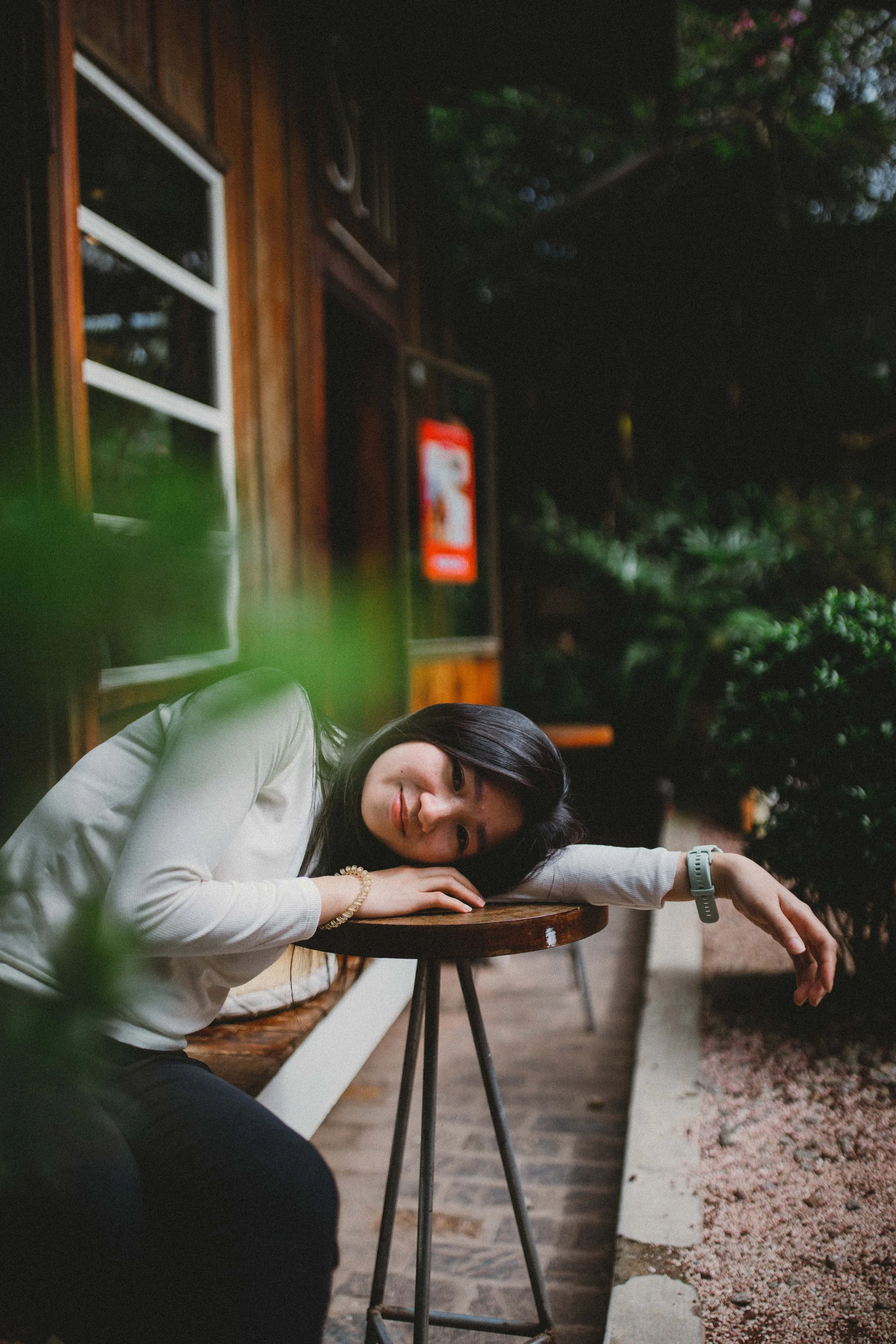 A woman with black hair wearing a white long-sleeve shirt and a smartwatch resting her head and arm on a round wooden table outdoors, with a background of greenery and a wooden building.