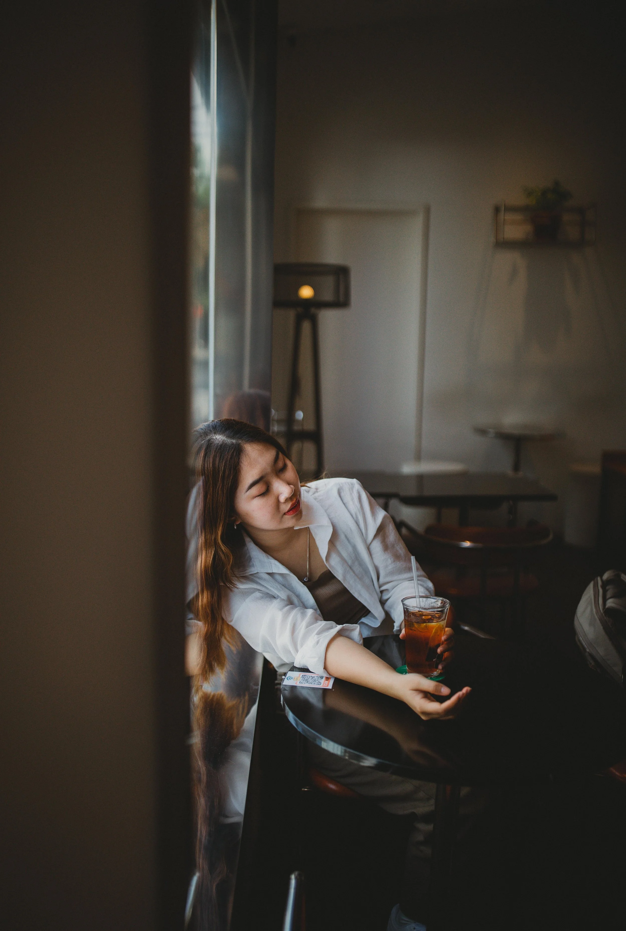 A young woman with long, wavy brown hair sitting at a black table in a cafe, holding a glass of iced tea with a straw, with her eyes closed, wearing a white shirt and a necklace.