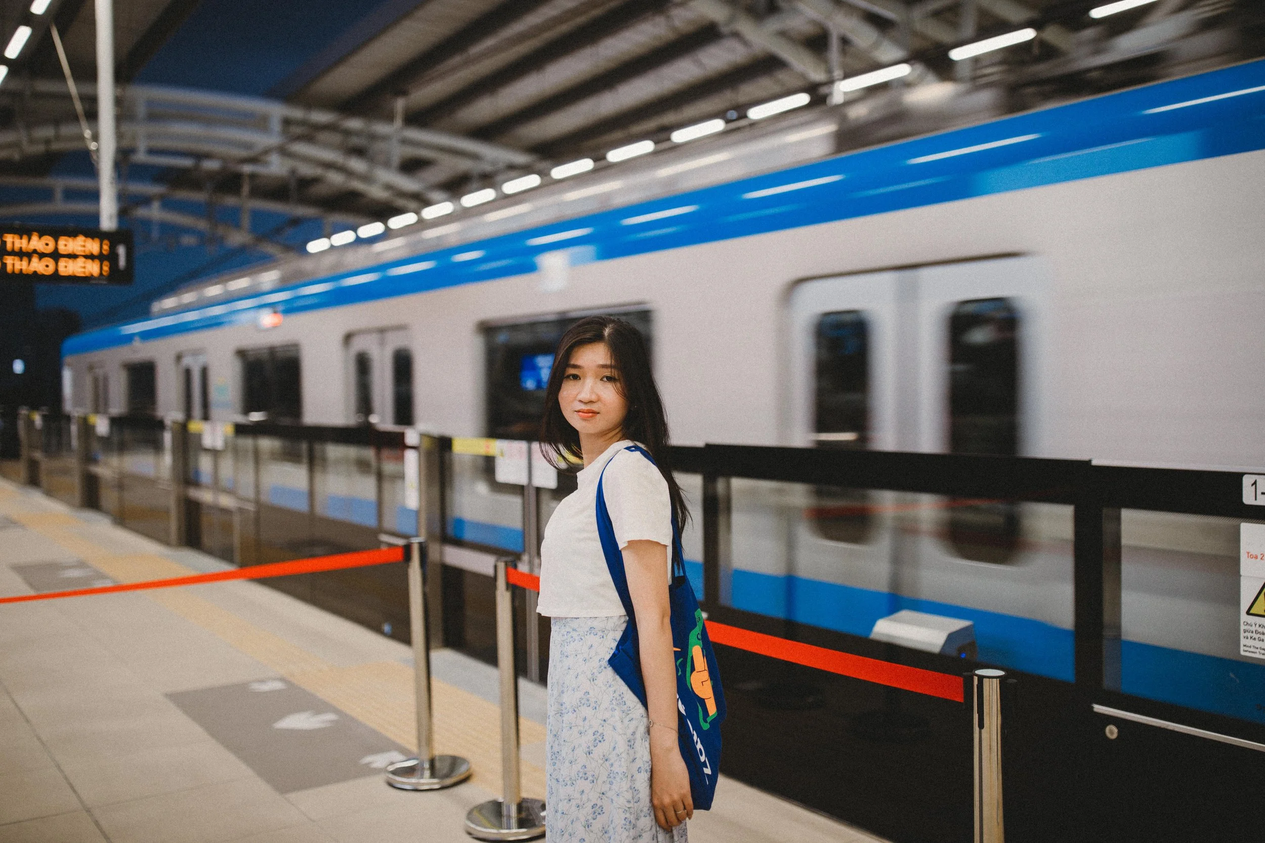 A young woman standing on a train station platform with a train arriving in the background at night.