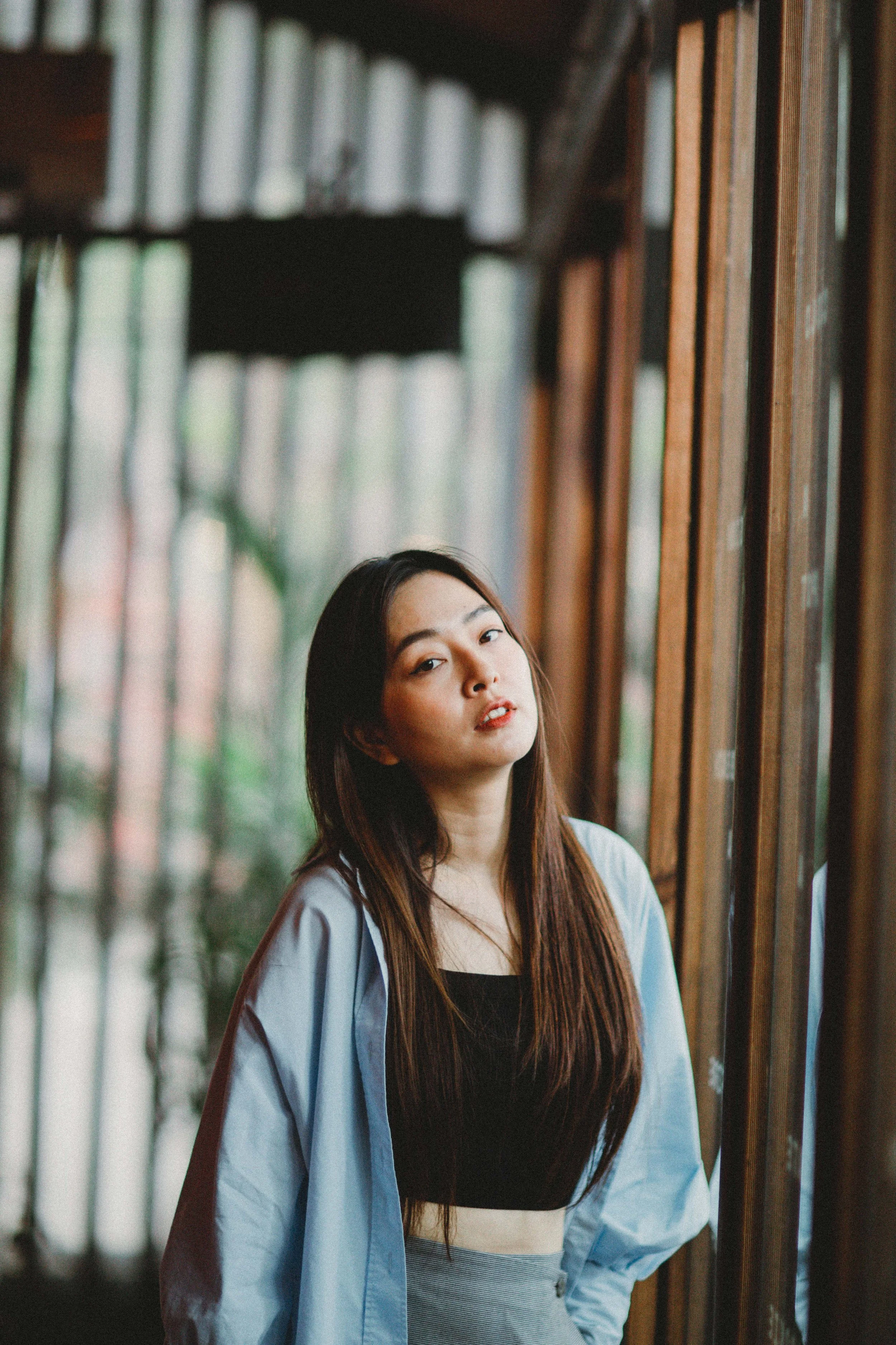 A young woman with long dark hair leaning against a wooden wall, looking at the camera with a neutral expression, standing indoors near large windows.