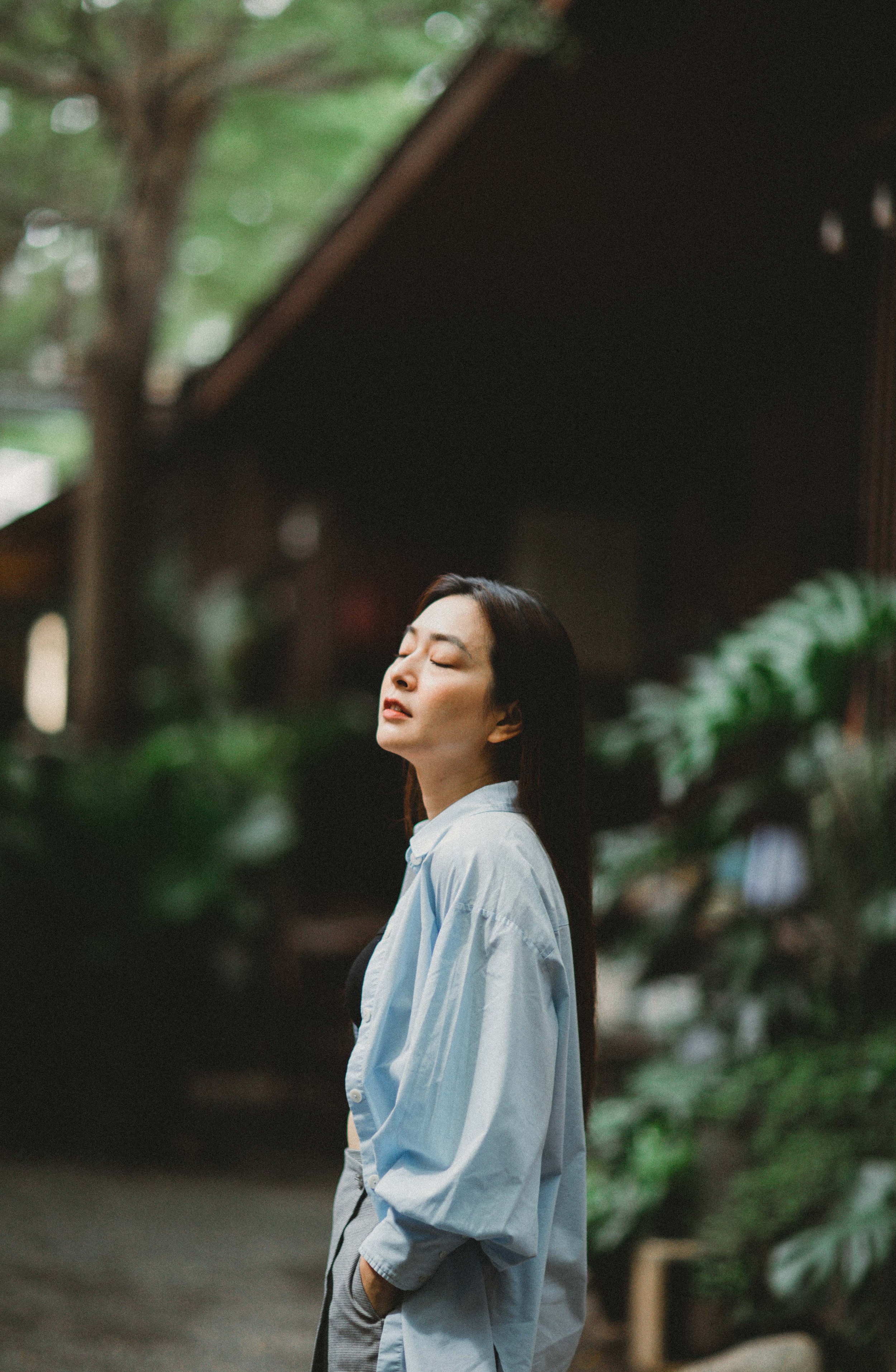 A woman with long dark hair, eyes closed, standing outdoors among greenery, wearing a light blue shirt.