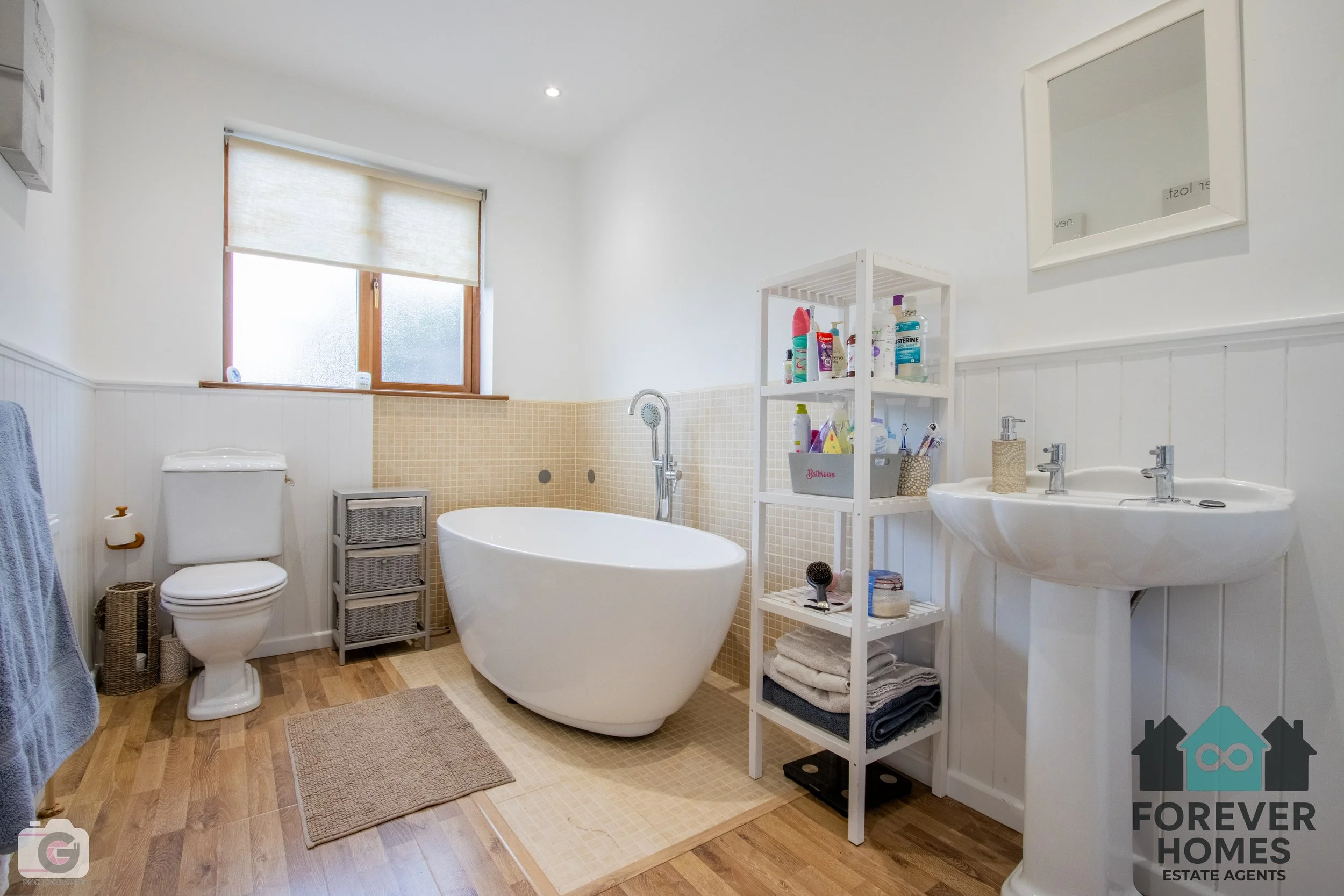 Bright bathroom with a white toilet, a white bathtub, a double-sink vanity, a white storage shelf with toiletries, a window with a beige blind, and wood-like flooring.