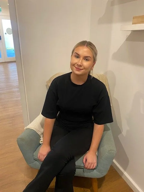 A young woman with shoulder-length brown hair and fair skin, wearing a black long-sleeve shirt and a delicate necklace, is sitting on a upholstered chair with a cream-colored blanket behind her, in a room with plain white walls.