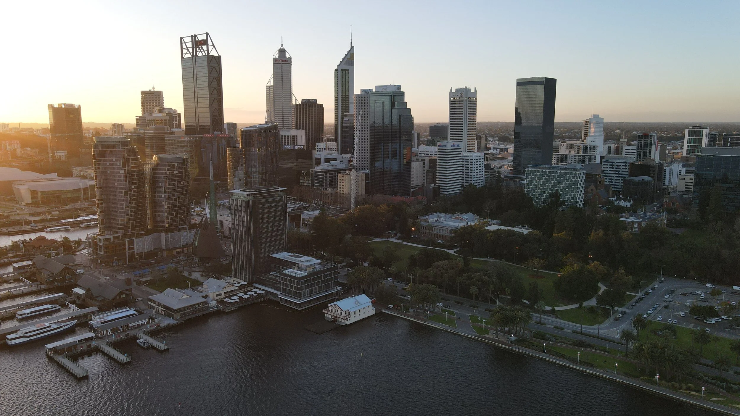 Aerial view of a cityscape with modern skyscrapers, waterfront buildings, and a park by the river at sunset.