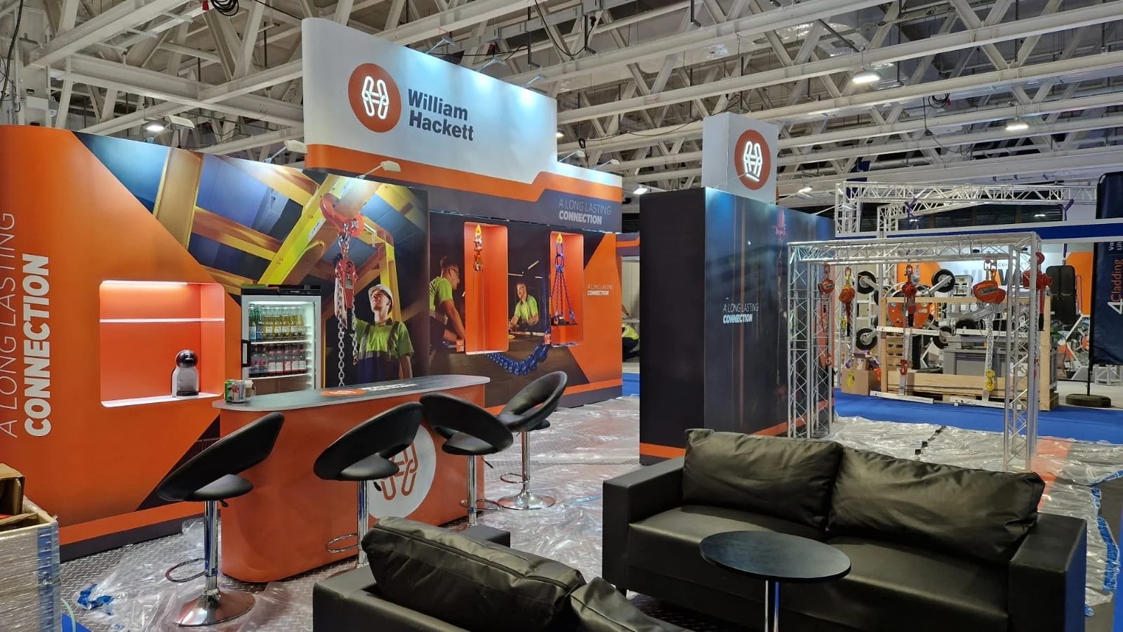 Trade show booth for William Hackett featuring orange and black branding, industrial lifting equipment, and seating area with black leather couches and a small table.