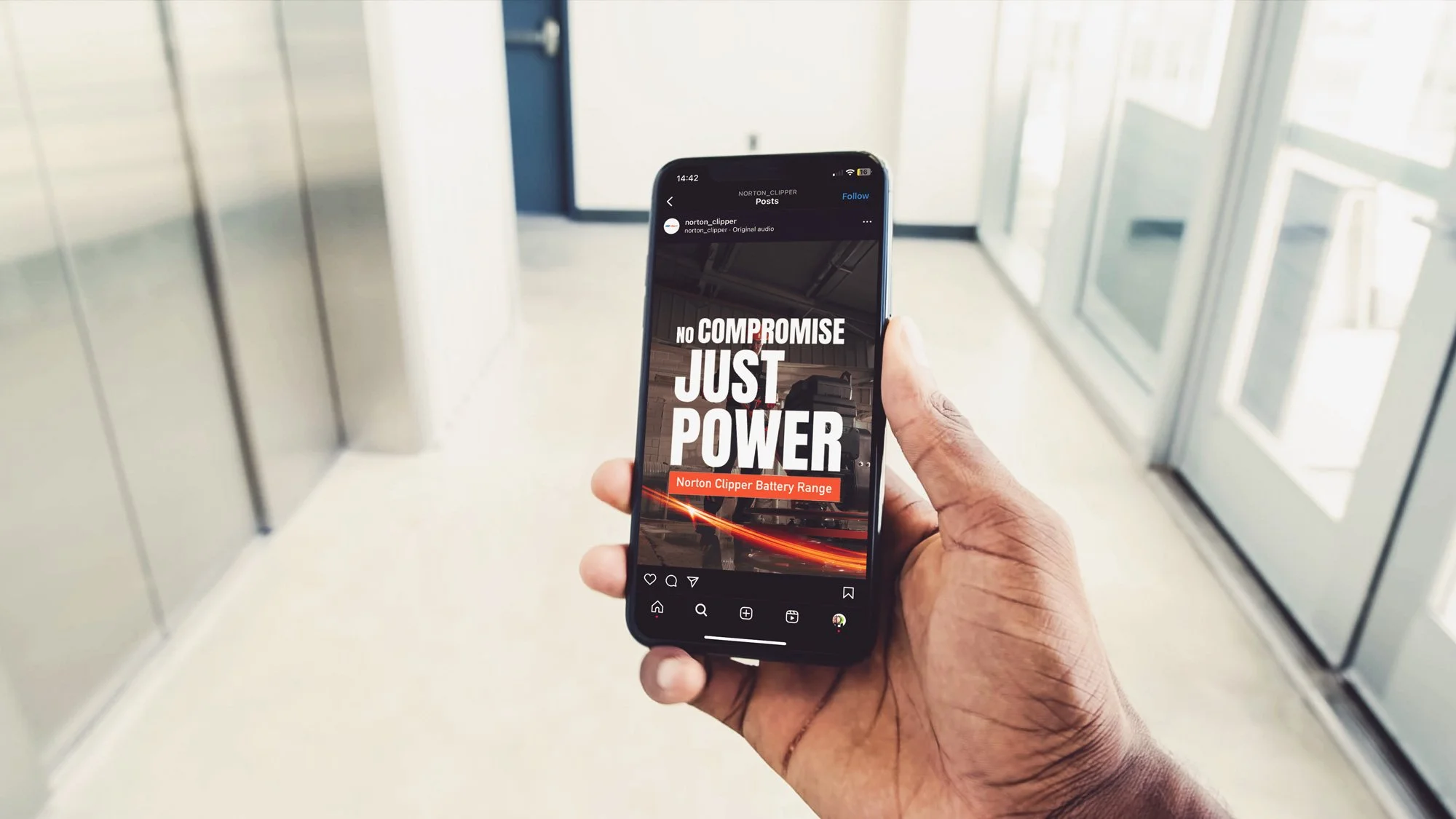 Person holding a smartphone displaying a graphic with the text 'NO COMPROMISE JUST POWER' and 'Norton Clipper Battery Range' in an indoor setting with an elevator.