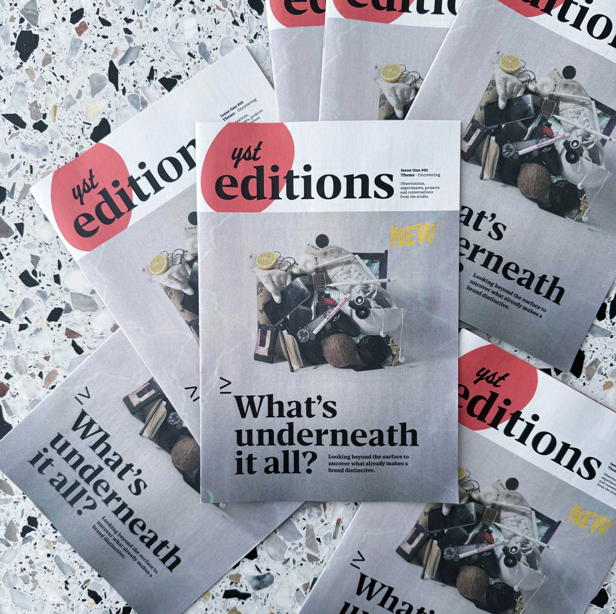 Several copies of a magazine titled 'yst editions' are spread on a surface with a terrazzo pattern. The magazines feature a cover with various objects including a lemon, a book, a smartphone, a gaming controller, a pair of glasses, and a microphone.