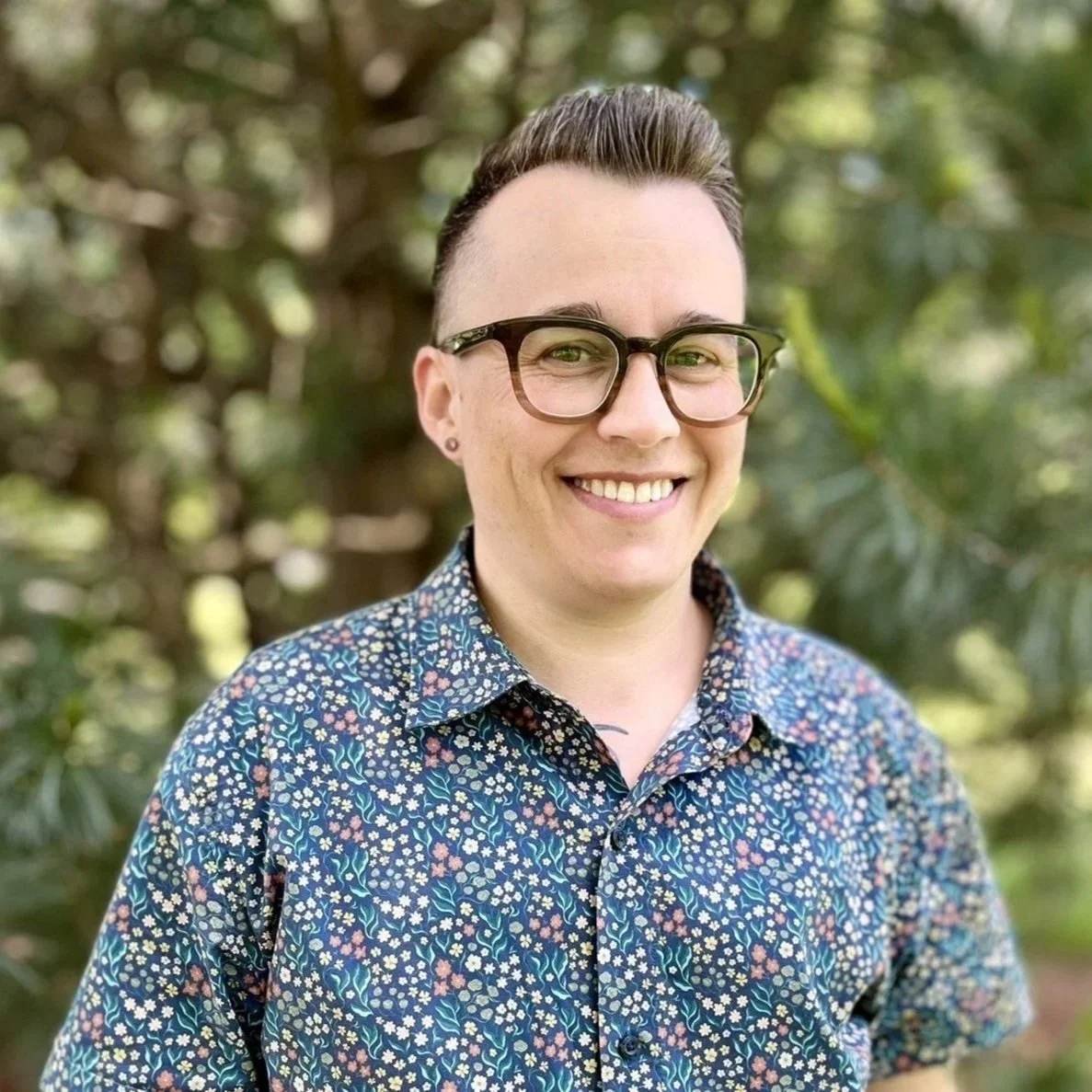 A person with short hair, glasses, and earrings, smiling outdoors with greenery in the background, wearing a blue floral patterned shirt.