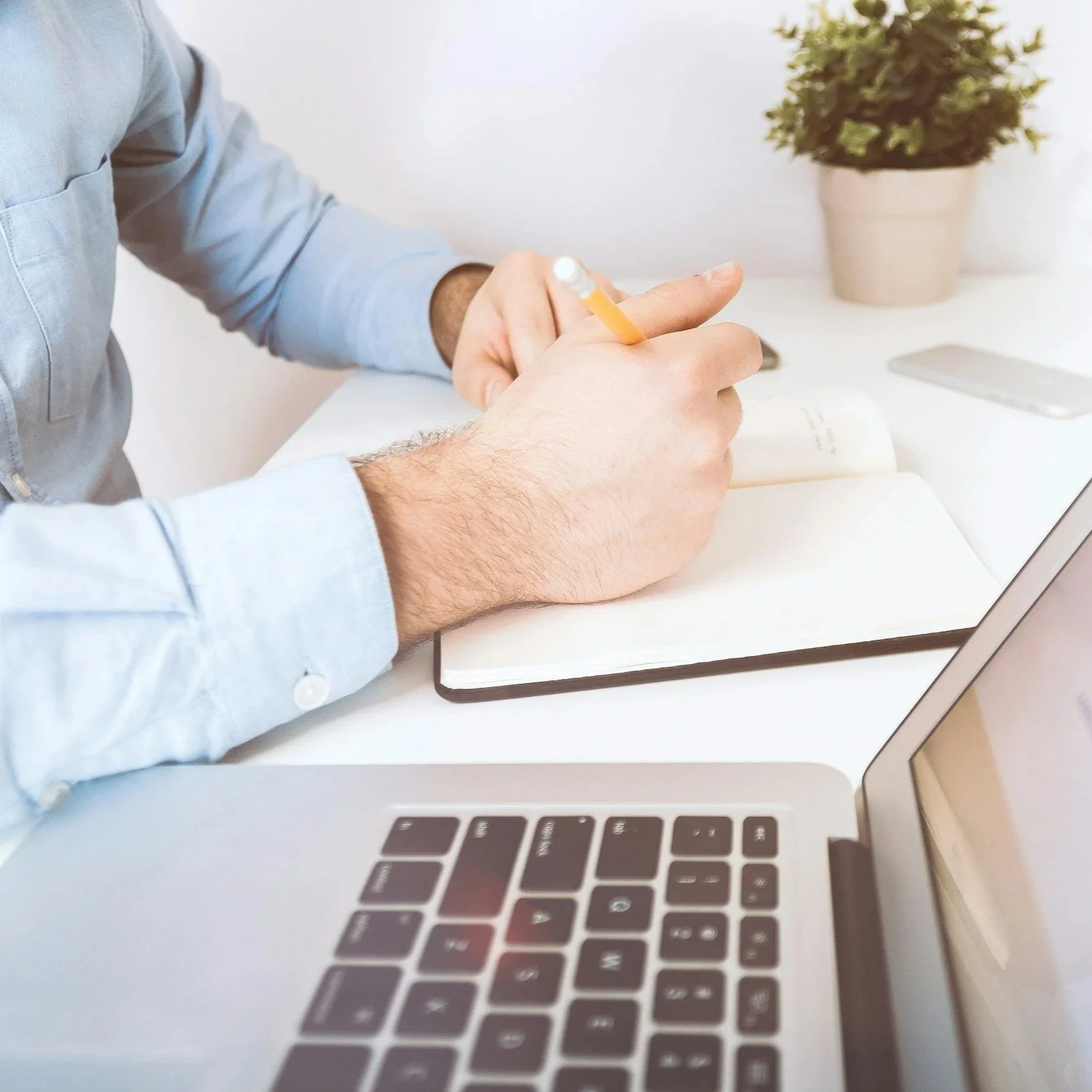 A person taking notes with a pencil in a notebook at a desk that has a laptop, a potted plant, and a smartphone.