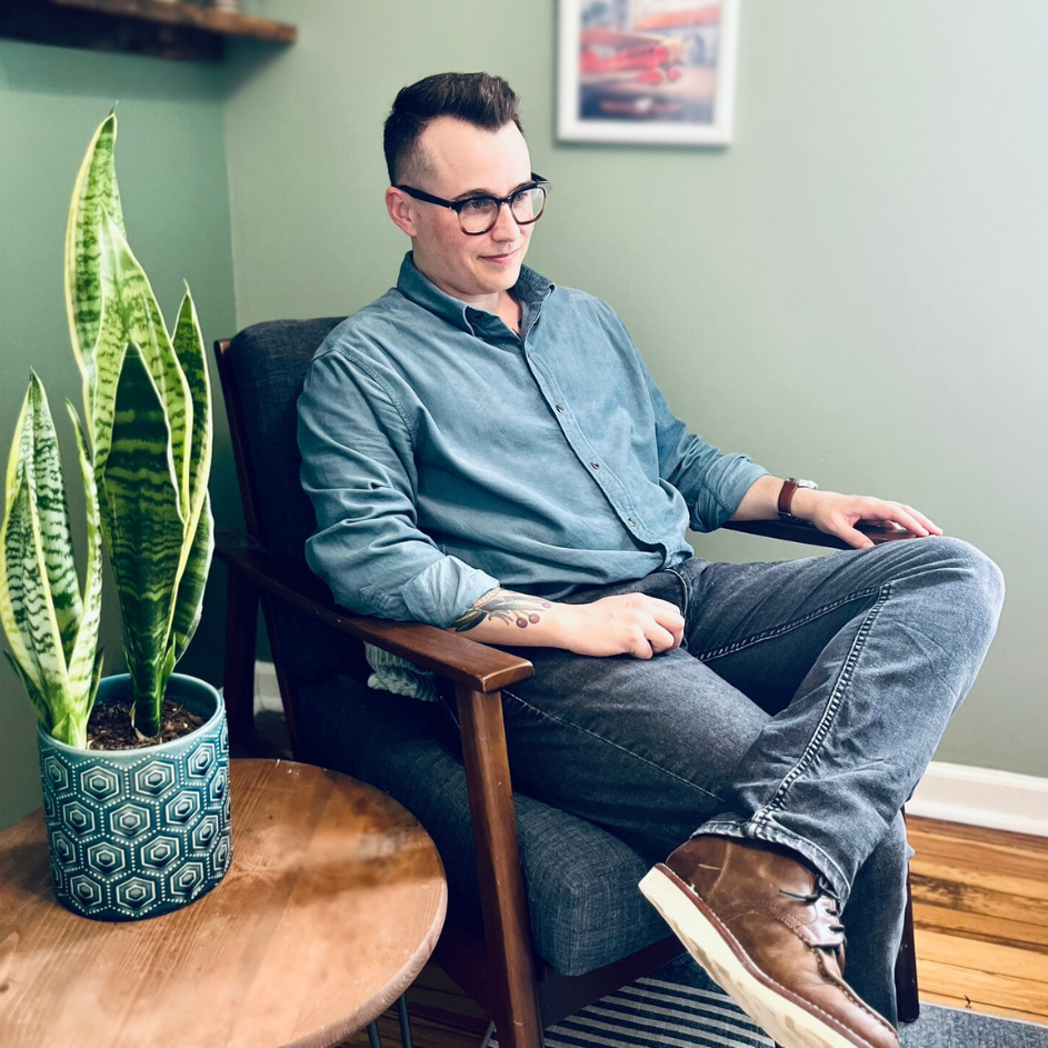 A person sitting in a chair in a blue shirt, jeans, and brown boots.They are wearing glasses and sitting next to a table with a plant on it.