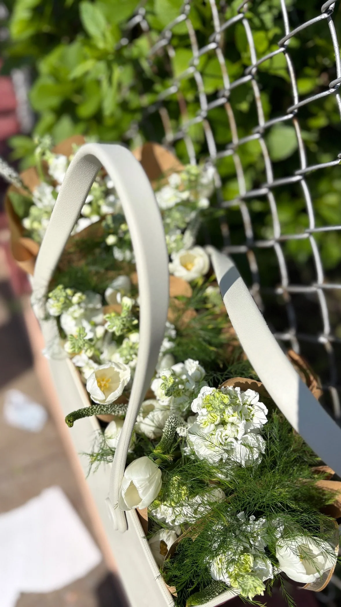 White and green floral bouquet in a tote bag by a chain-link fence with greenery.