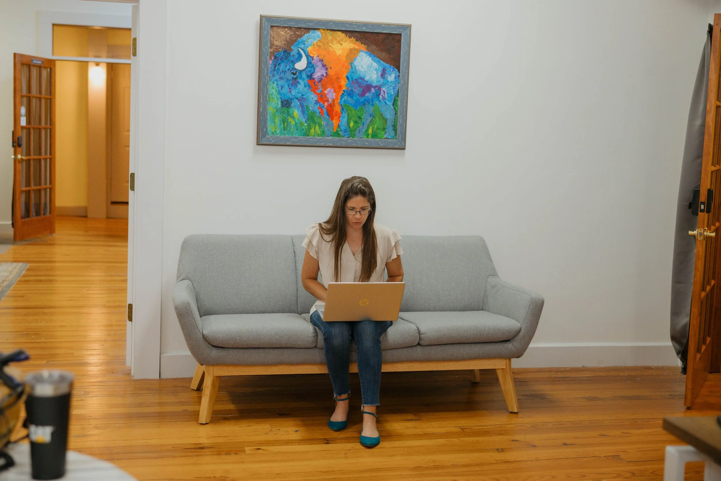 A young woman sitting on a gray couch in a living room, working on a silver laptop, with a plaid blanket behind her. In front, there is a white coffee table holding magazines. The wall behind her has a large framed quote and a smaller framed photo. The room has wood flooring and a white ceiling with tiled pattern and a rectangular light fixture.