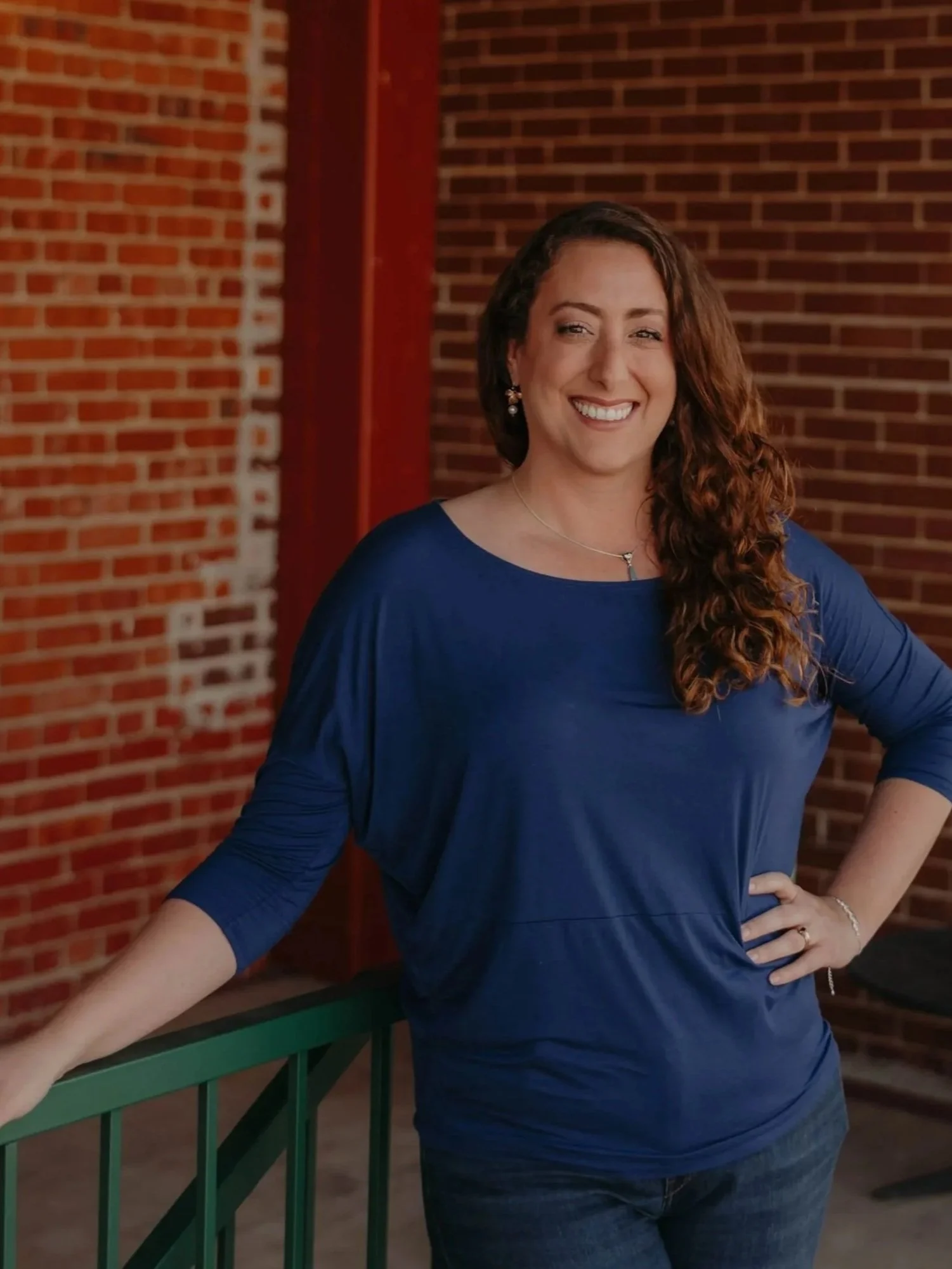 A smiling woman with curly brown hair, wearing a blue top and jeans, standing outdoors with a brick wall background, leaning on a green railing.