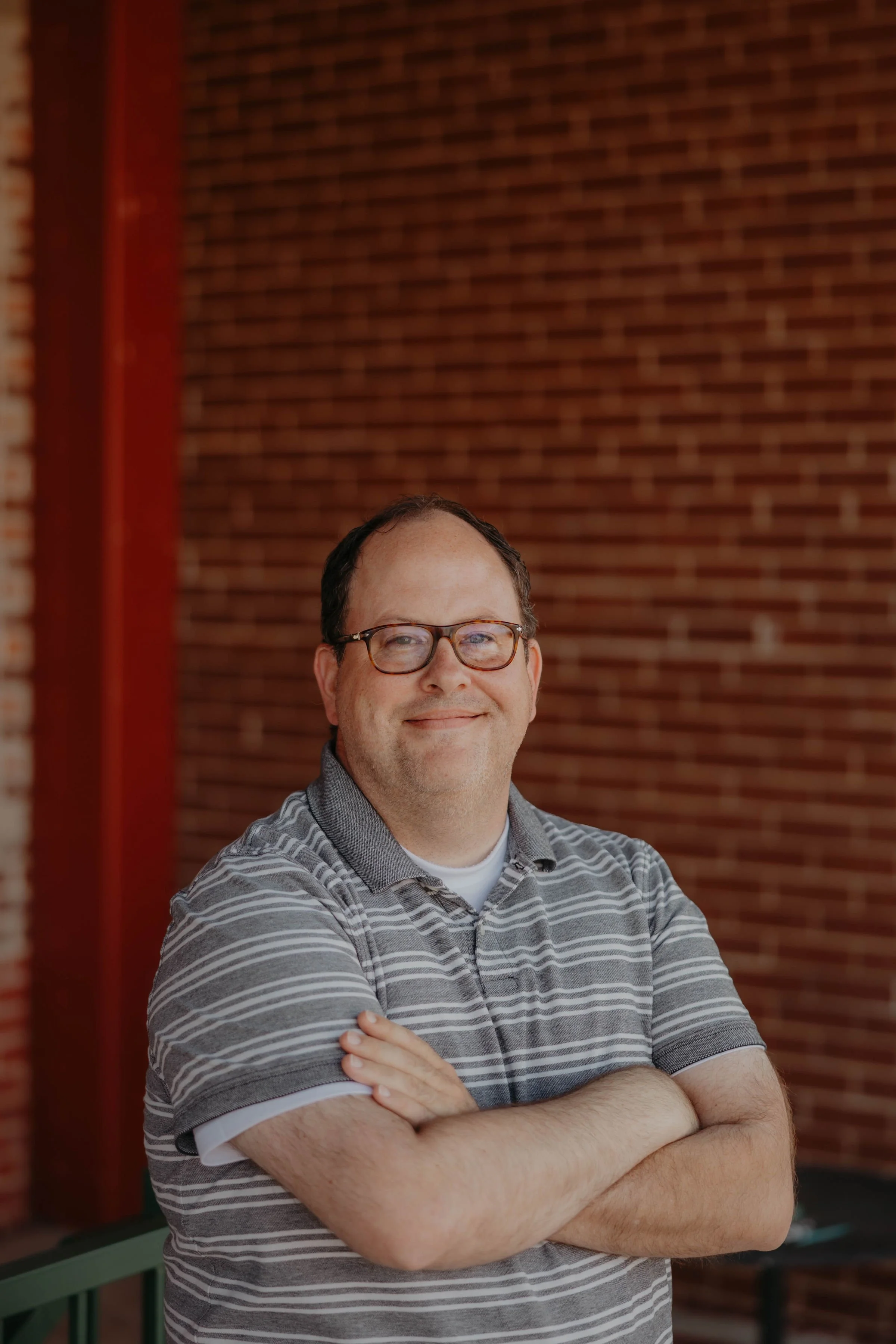 A man with glasses wearing a gray striped polo shirt, standing with arms crossed, smiling, against a brick wall background.