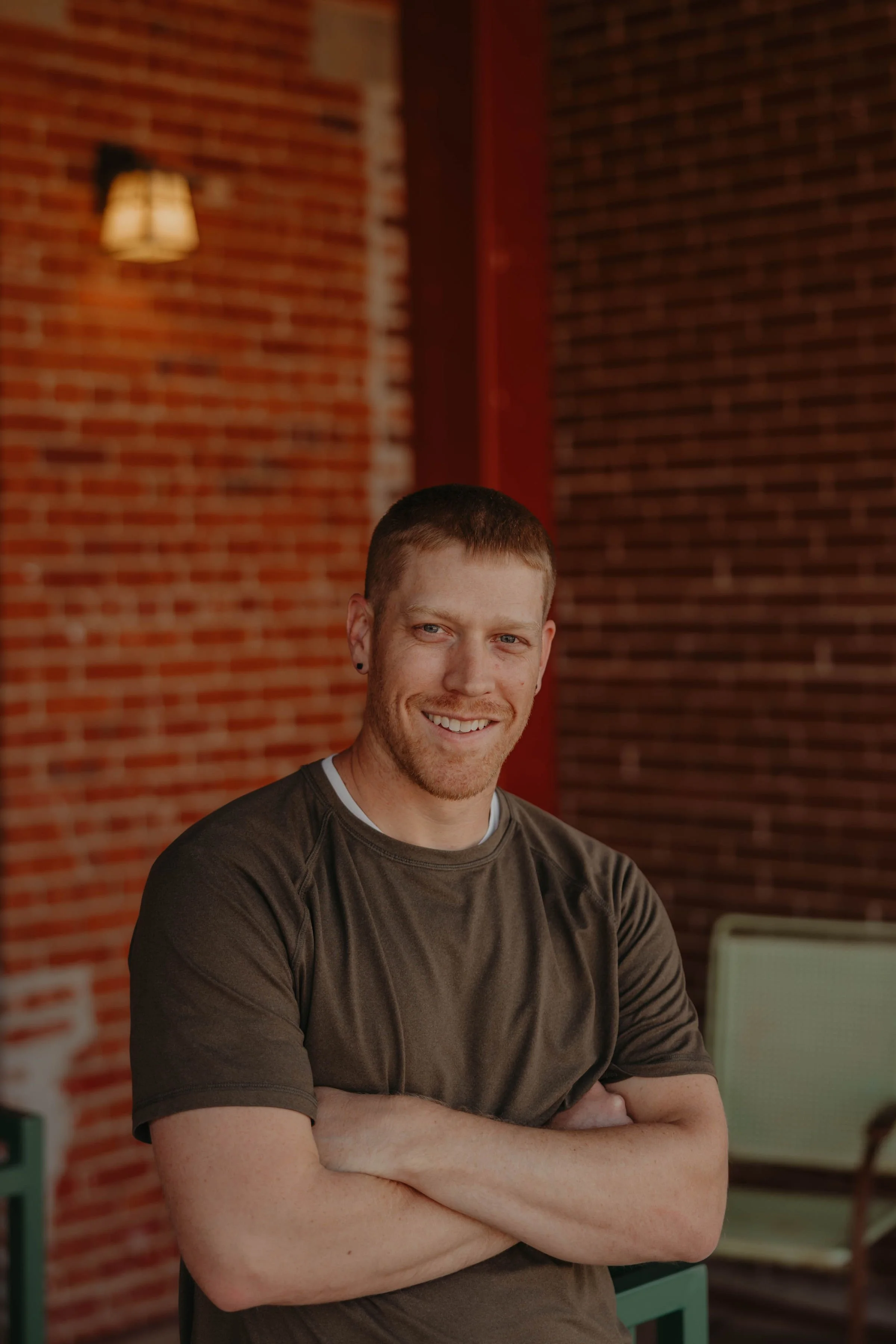 A smiling man with short red hair and a beard, crossing his arms, in front of a brick wall with warm lighting.