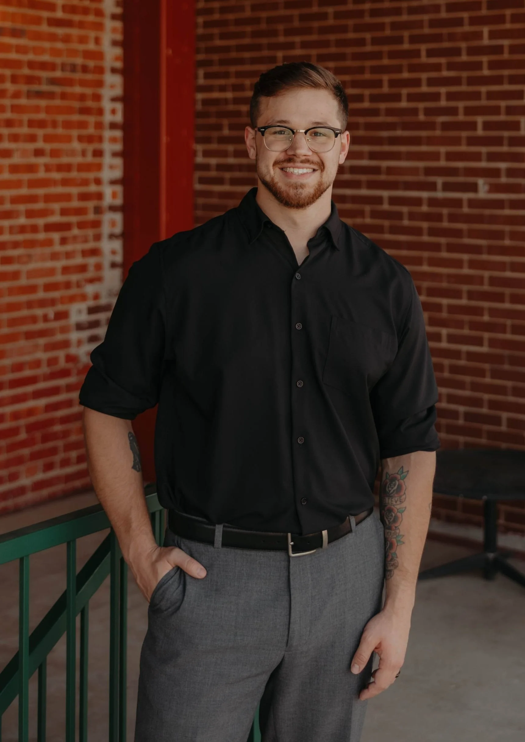 Young man with glasses, beard, and tattoos, wearing a black shirt and gray pants, smiling and standing outdoors against a brick wall.