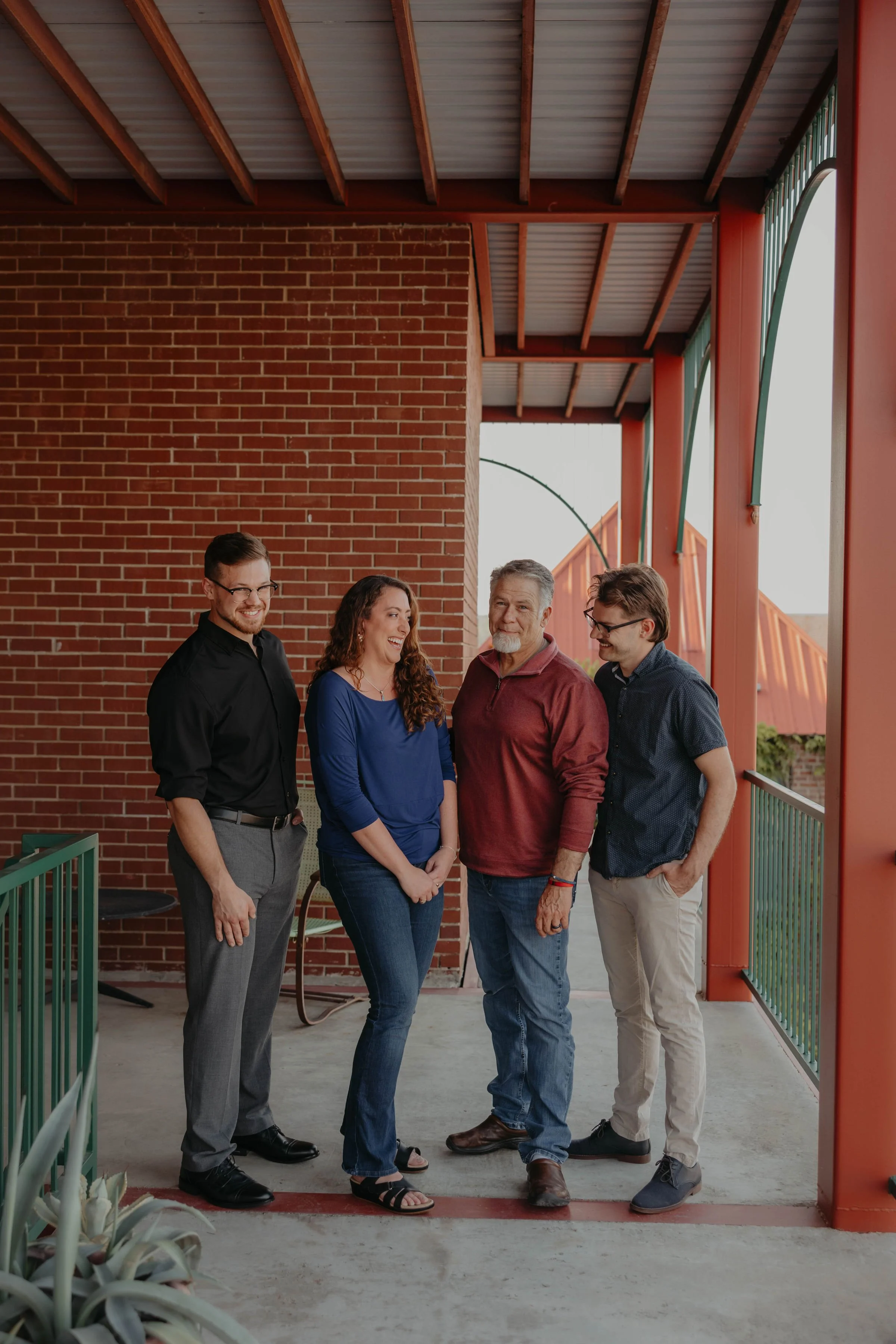 Four people standing together on a balcony with a brick wall and metal railing, smiling and talking.