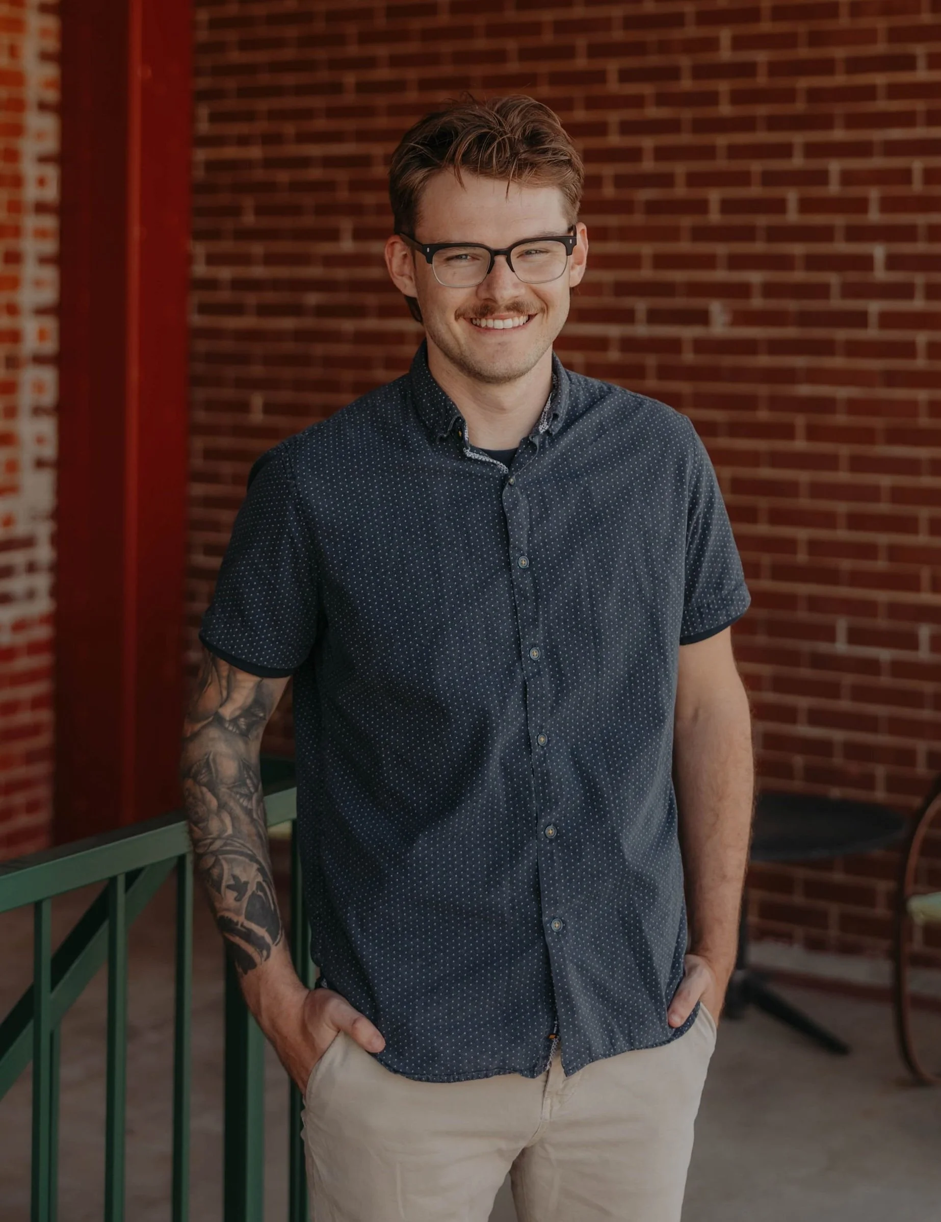 A young man with glasses, a mustache, and tattoos on his right arm, smiling and standing outdoors next to a green railing against a brick wall background.