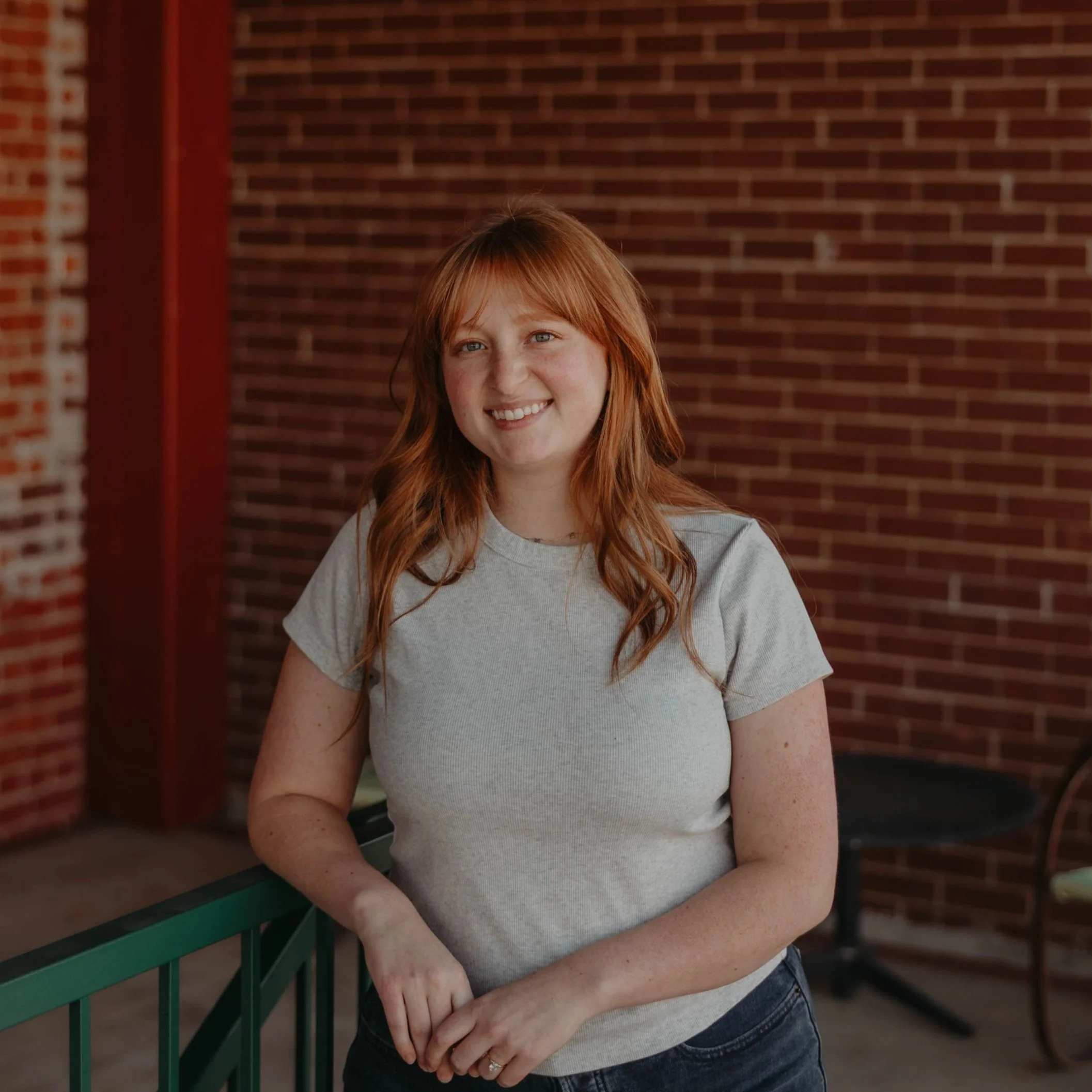 A young woman with red hair in a gray t-shirt standing outdoors near a brick wall, smiling, with green railing in front.
