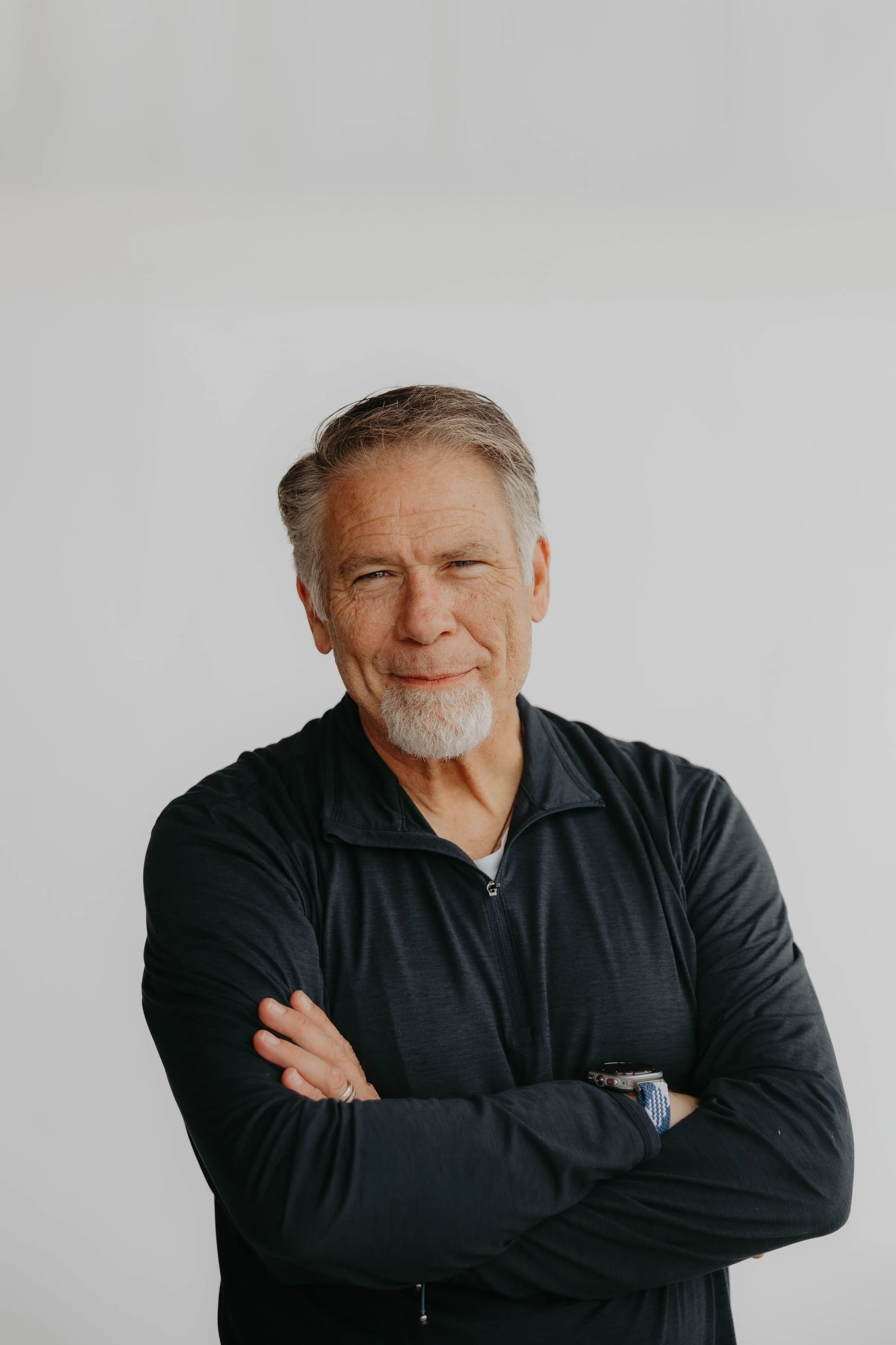 Smiling older man with gray hair and beard wearing a maroon pullover and blue jeans, leaning on a green railing against a red brick wall.