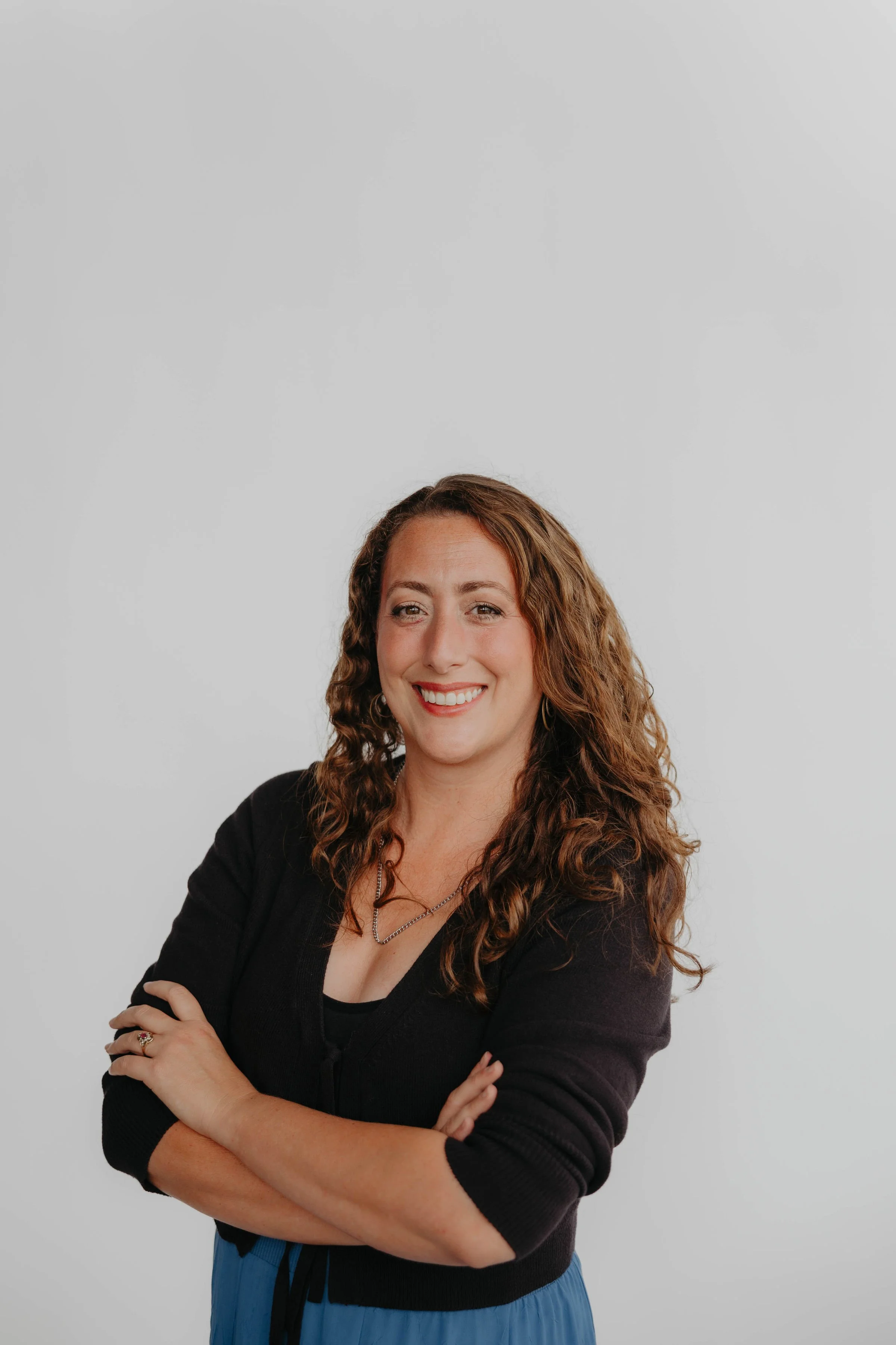 A smiling woman with curly brown hair, wearing a blue top and jeans, standing outdoors with a brick wall background, leaning on a green railing.