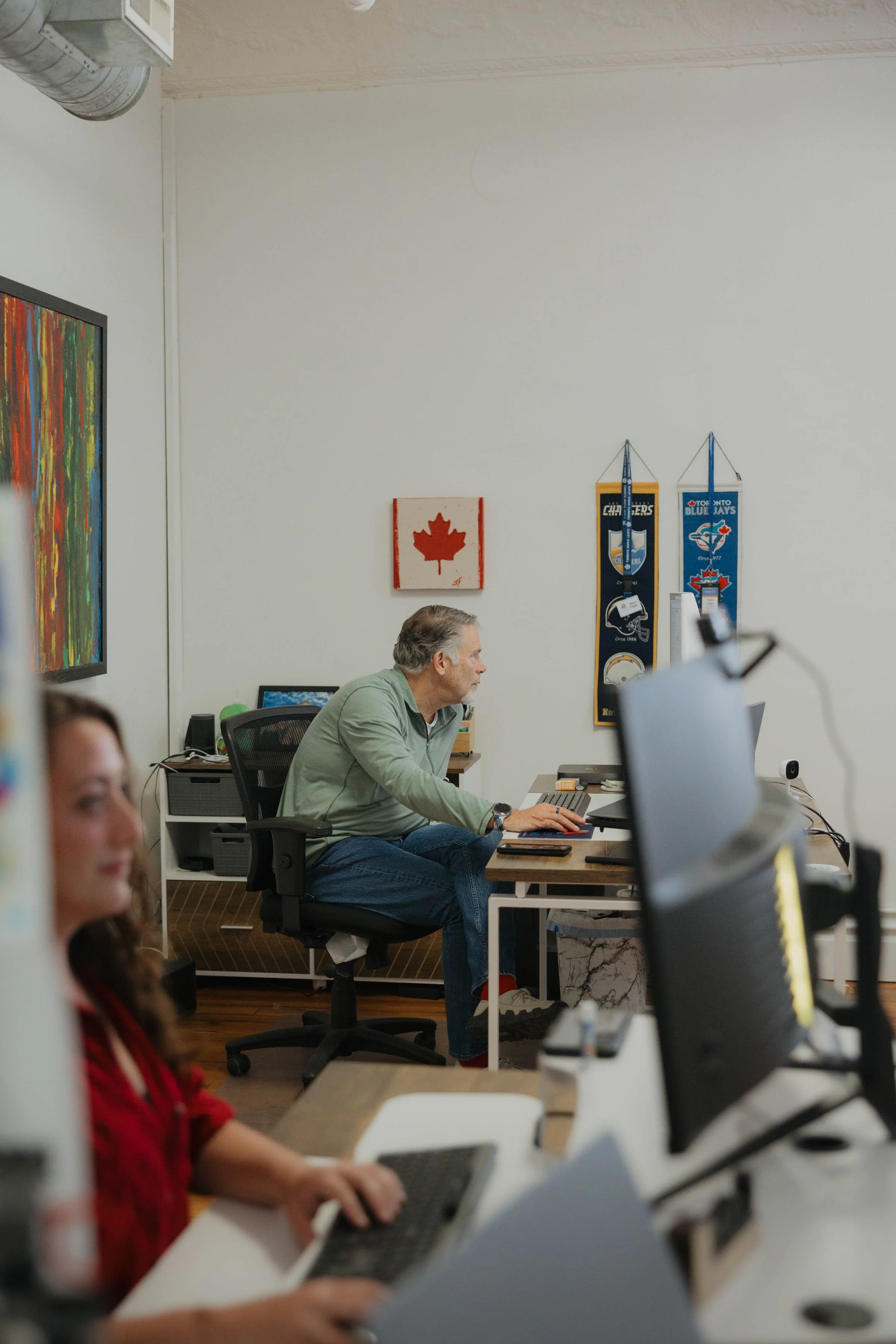 Two men working on an open computer tower at a white desk, with a monitor, tools, and a fire extinguisher nearby in an office with white walls and framed pictures.