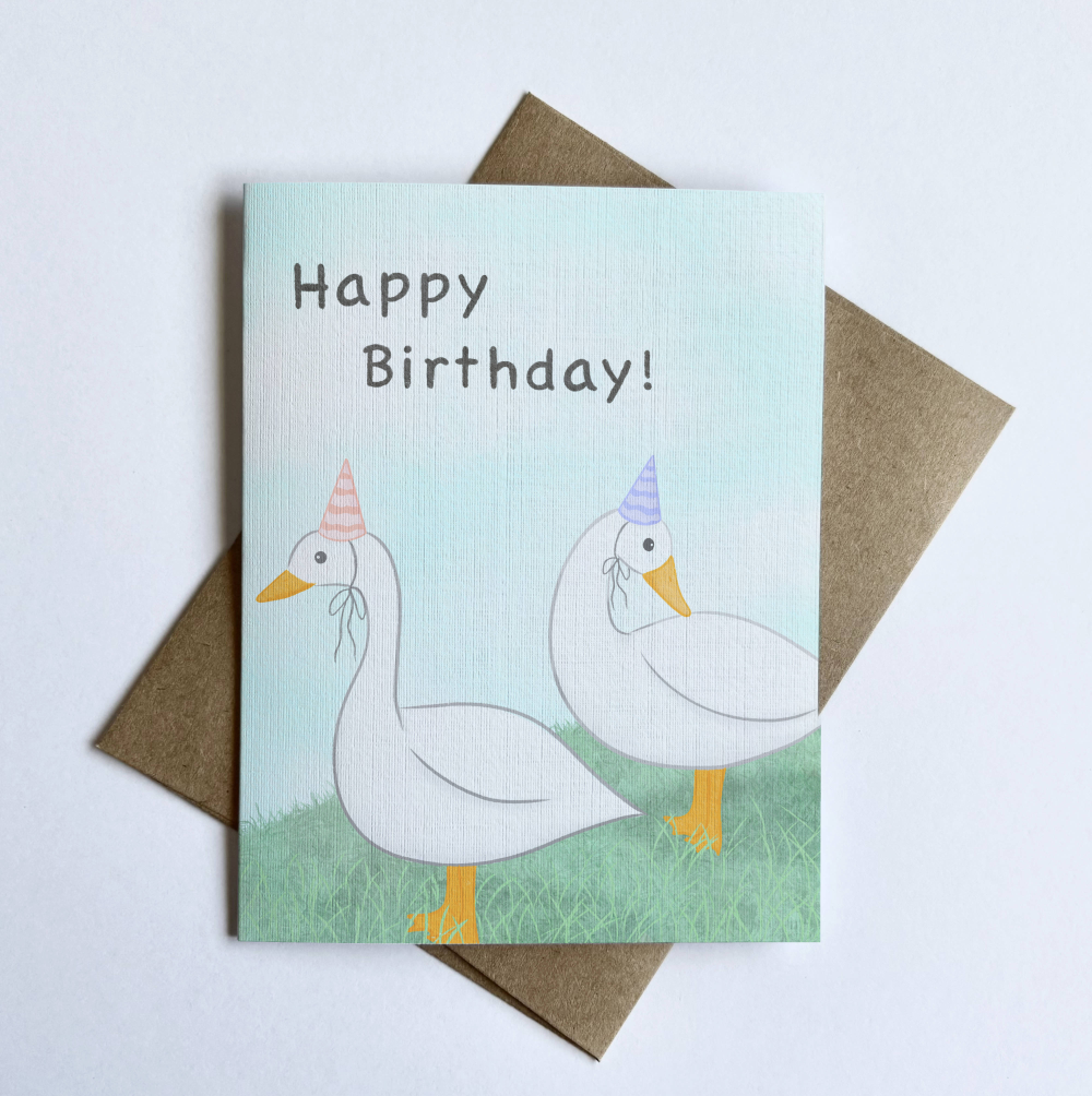A closeup of an illustration of ducks standing on grass wearing pastel pink and purple party hats against a soft blue cloudy background. The card text on the front says “Happy Birthday.”