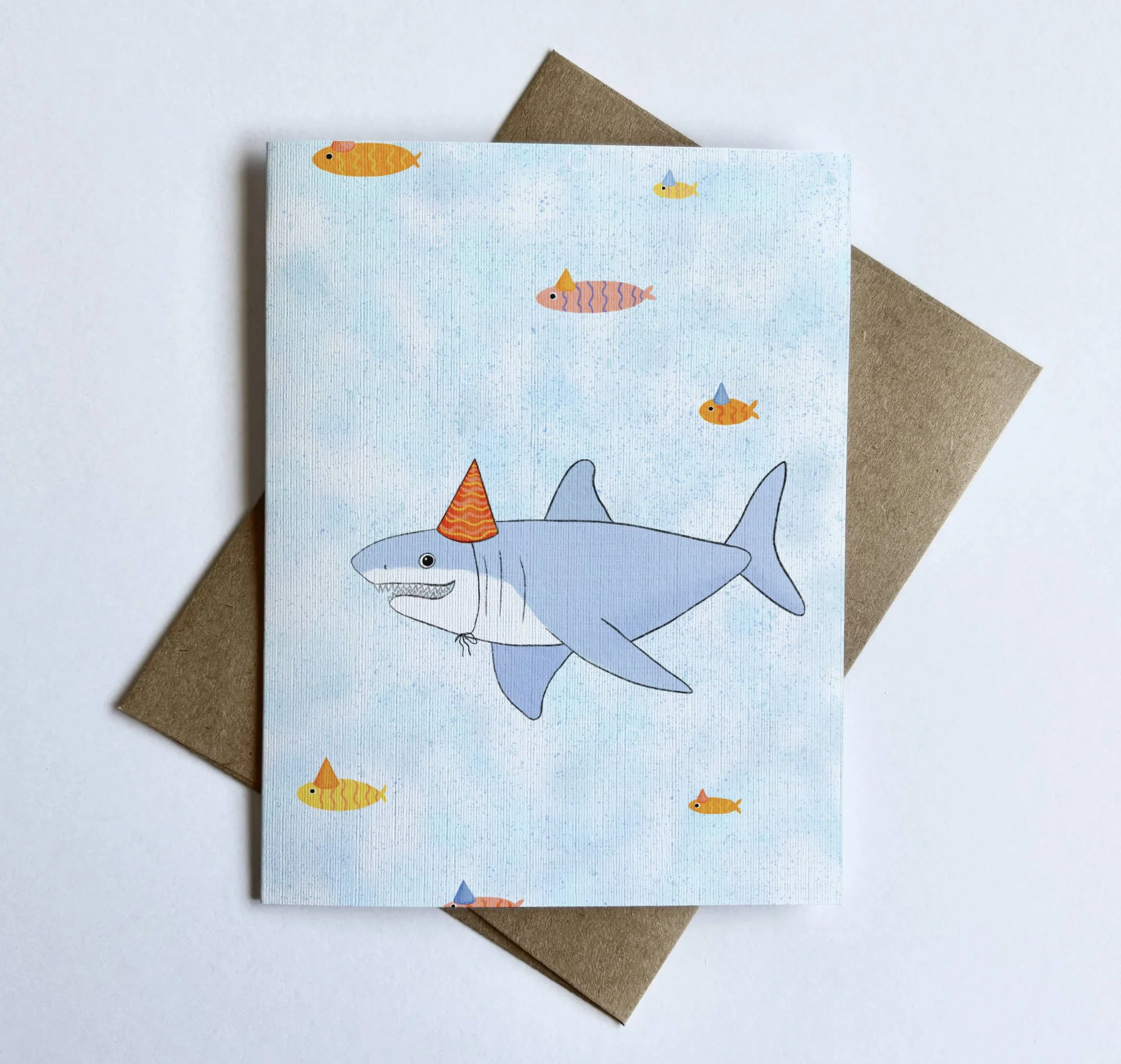 Closeup of a illustration of a humorous shark wearing a party hat, with tiny party-hat fish swimming in the background, on a blank linen textured card.