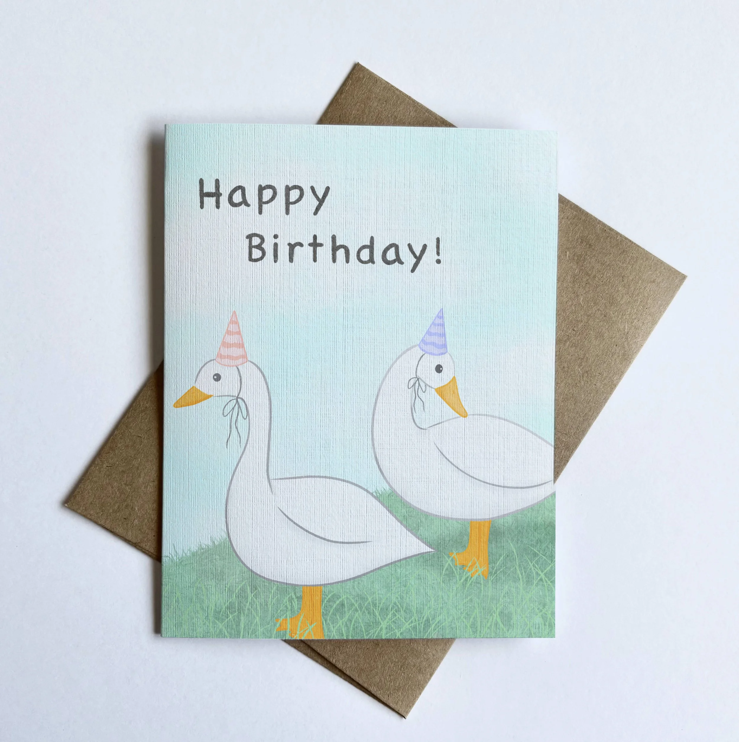 A closeup of an illustration of white ducks standing on grass wearing pastel pink and purple party hats against a soft blue cloudy background. The card text on the front says “Happy Birthday.”
