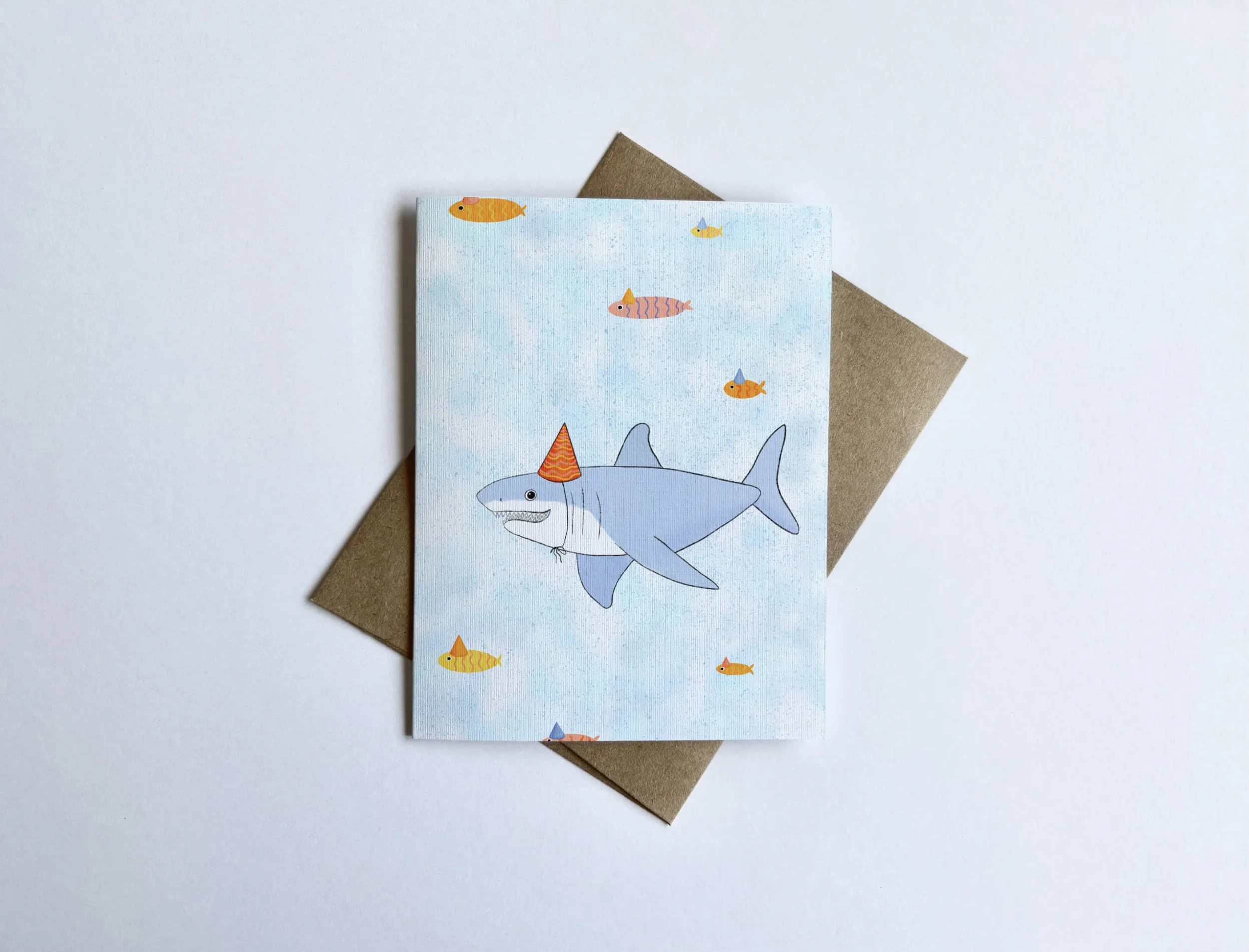 An illustration of a humorous shark wearing a party hat, with tiny party-hat fish swimming in the background, on a blank linen textured card.