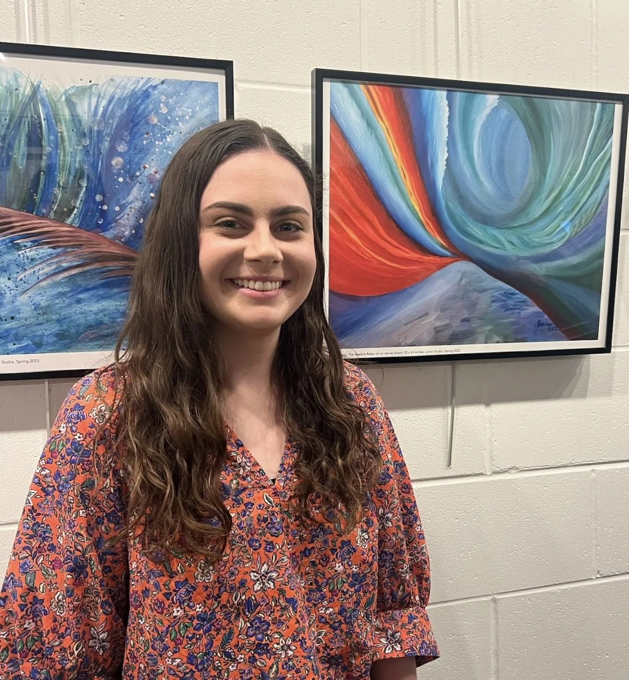 A headshot photo of the Artist, Writer, and Designer Jenna Baerg standing in front of prints of two of her abstract paintings. Jenna is smiling and has long wavy brown hair. Her painting, "The Healer's Robe" is in the background on the right.