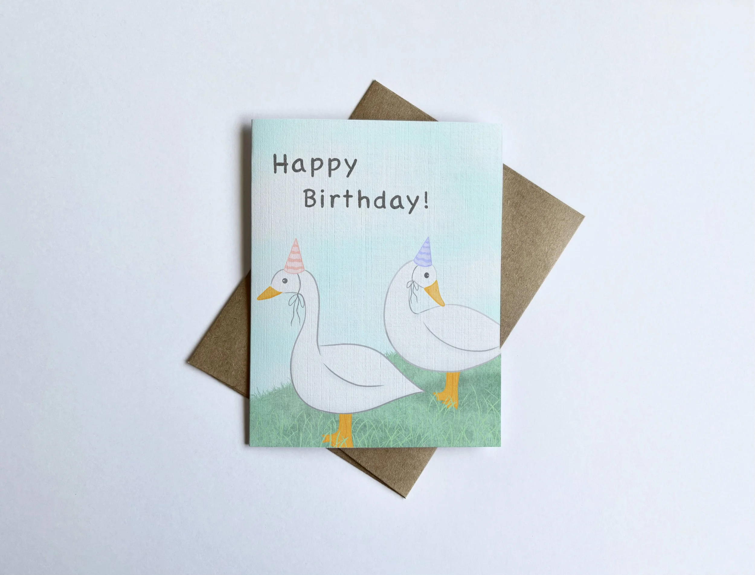 An illustration of ducks standing on grass wearing pastel pink and purple party hats against a soft blue cloudy background. The card text on the front says “Happy Birthday.”