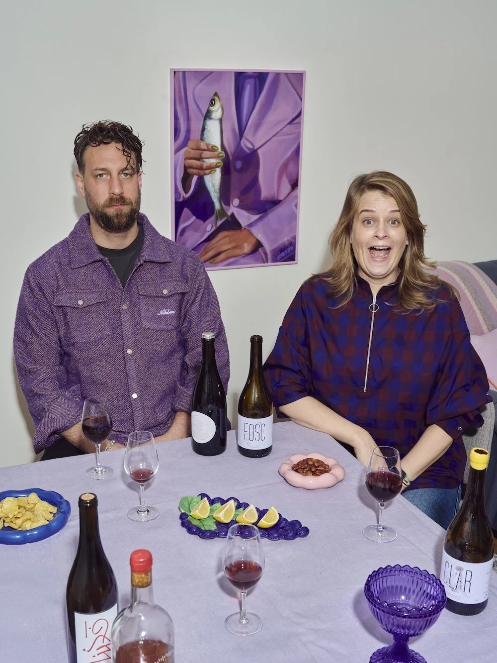 En kvinna och en man sitter vid ett middagsbord med dryck och snacks, ler och poserar för kameran. På väggen bakom dem finns en målning av en person som håller en fisk.