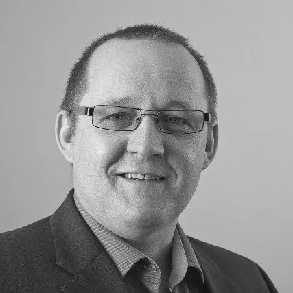 Black and white photo of a man with glasses, wearing a suit and collared shirt, smiling.