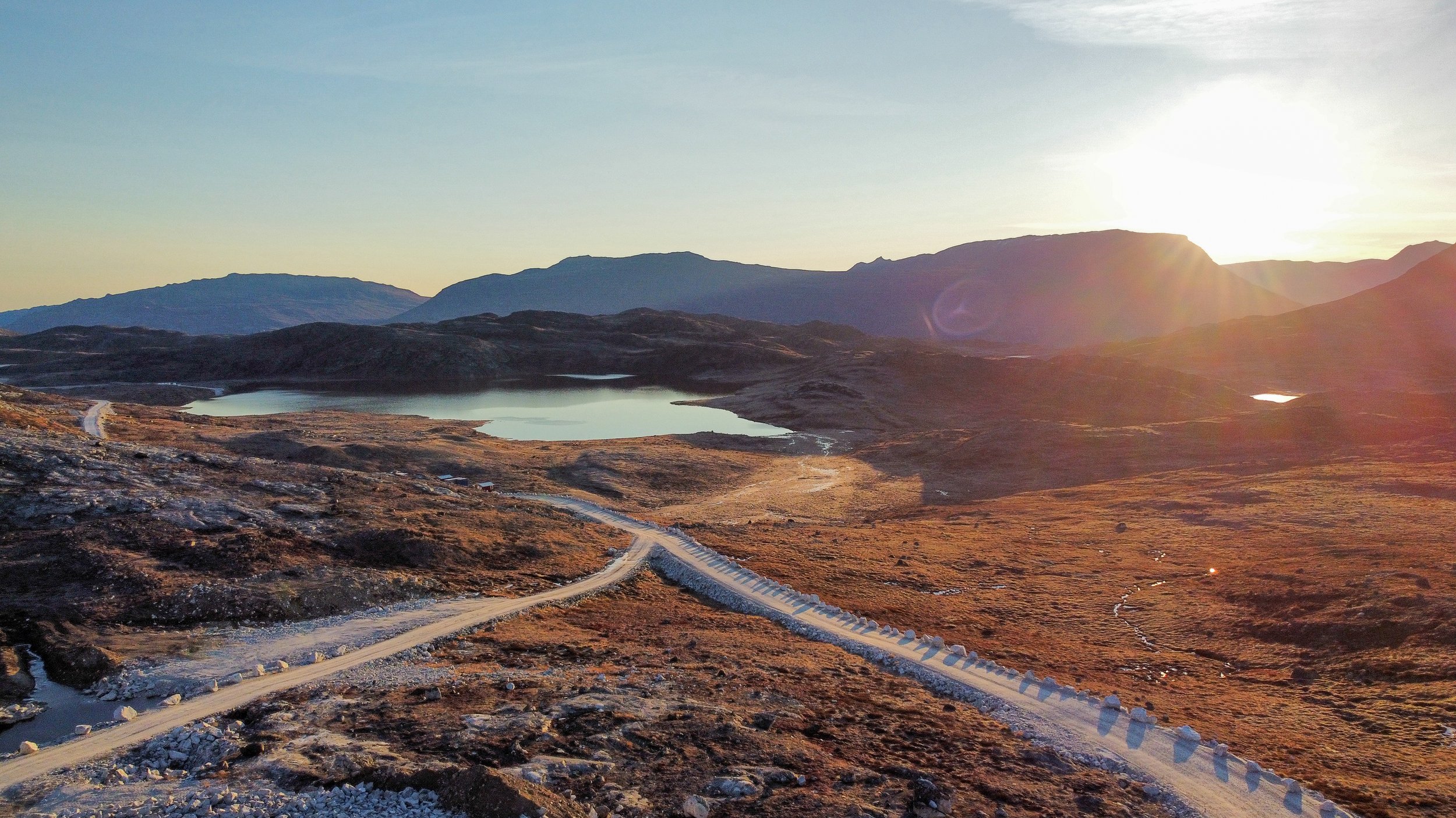 A dirt road winding through a mountainous landscape with small lakes, rocky terrain, and the sun setting behind distant mountains.